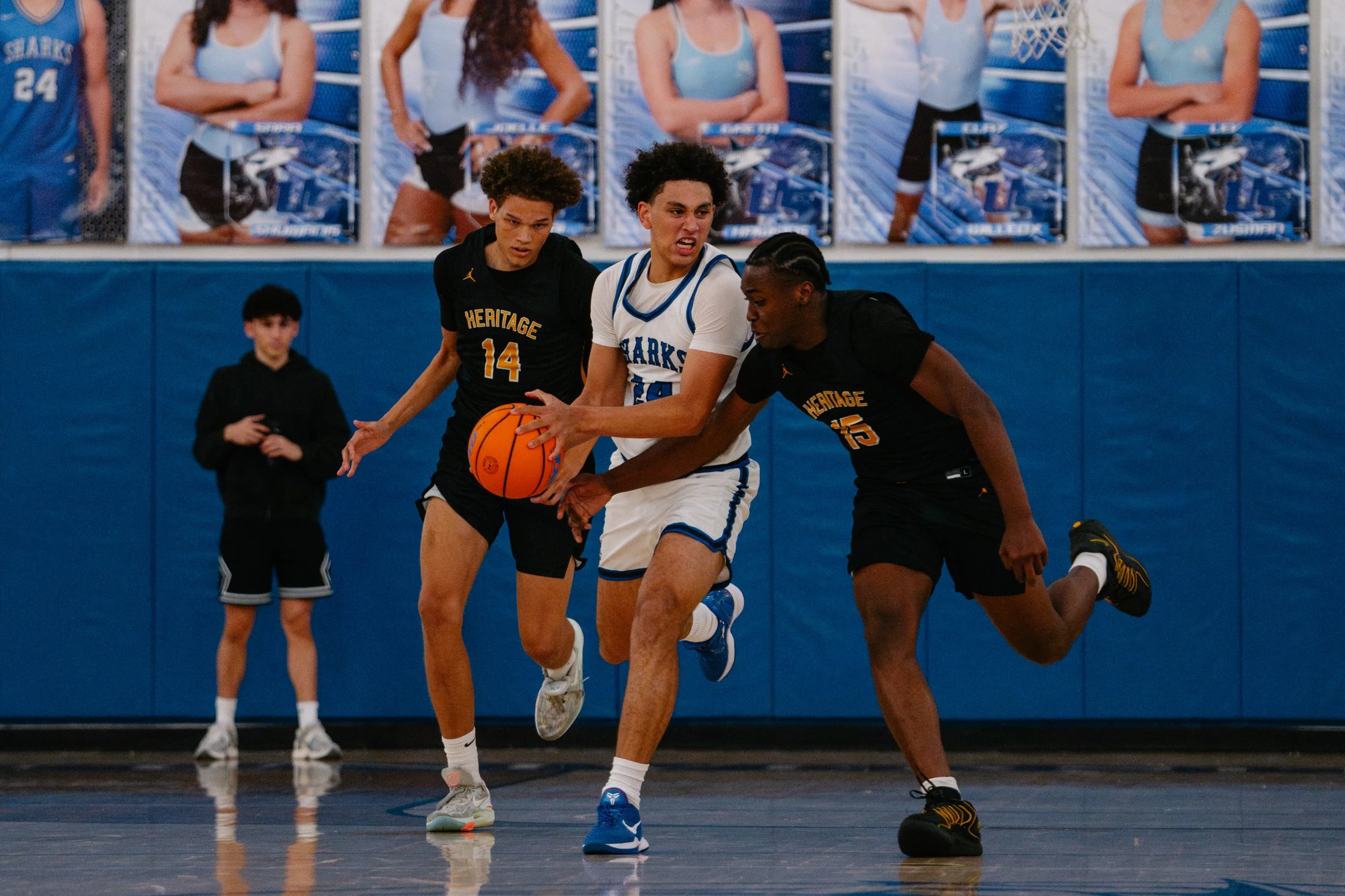 Two basketball players from Heritage High School in black uniforms and a player from a team in a white and blue uniform compete for possession of the basketball on a gymnasium court. One of the Heritage players is wearing number 14, the other is numb
