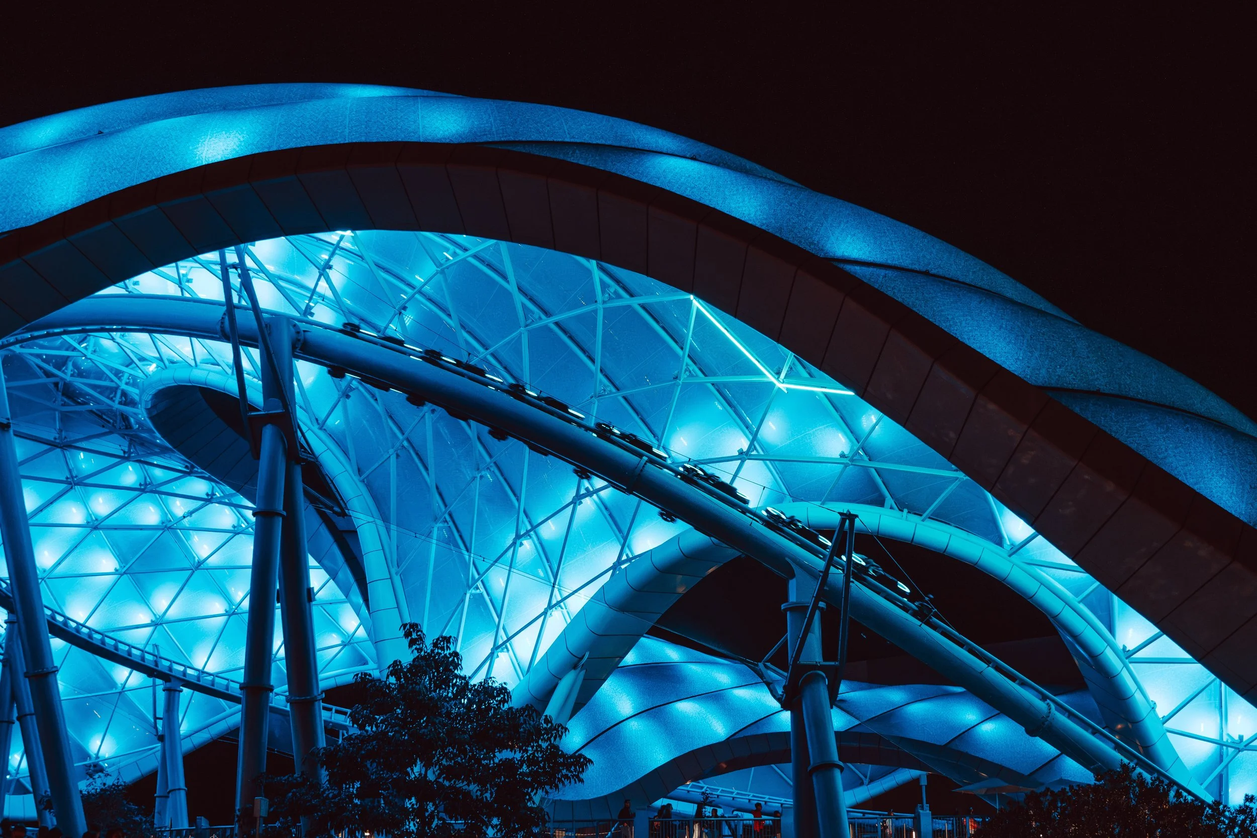 Night view of an illuminated futuristic architectural structure with curved metallic surfaces and a glass dome, lit in blue.