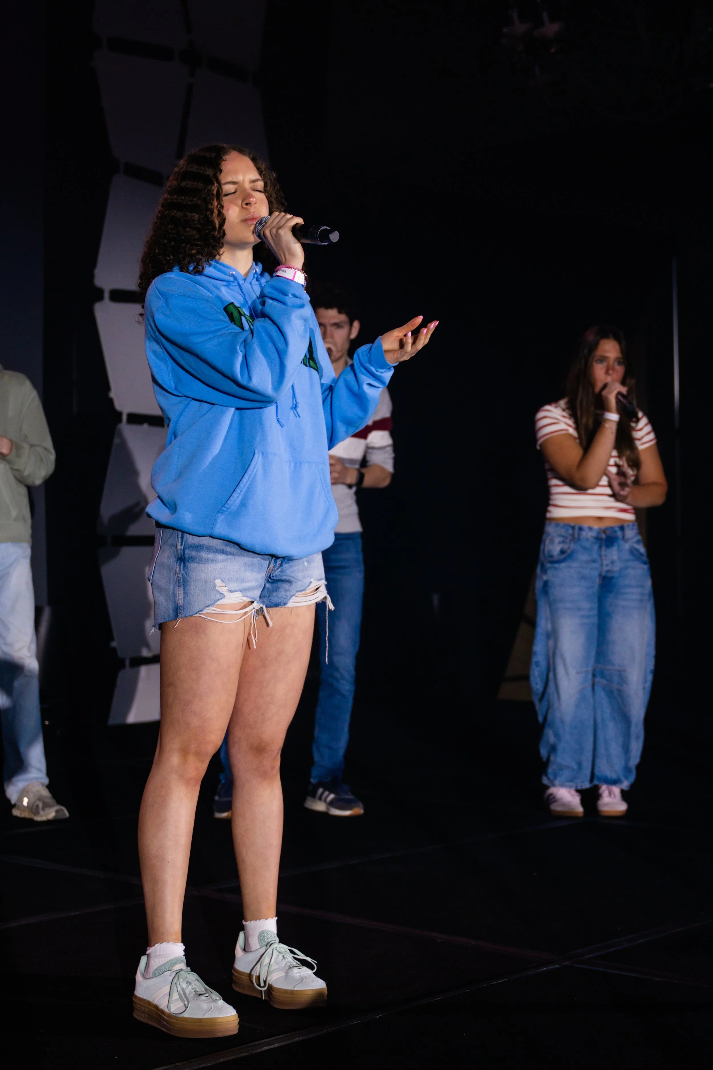 A group of young people on a stage, with a woman in a blue hoodie and ripped shorts singing into a microphone, and others standing in the background.