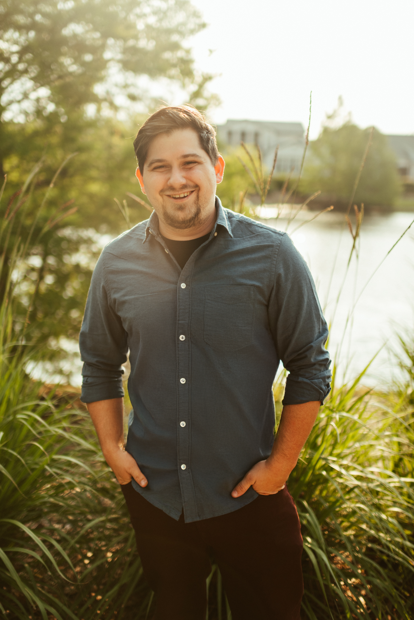 A man smiling outdoors near a body of water and greenery, with a building in the background during sunset.