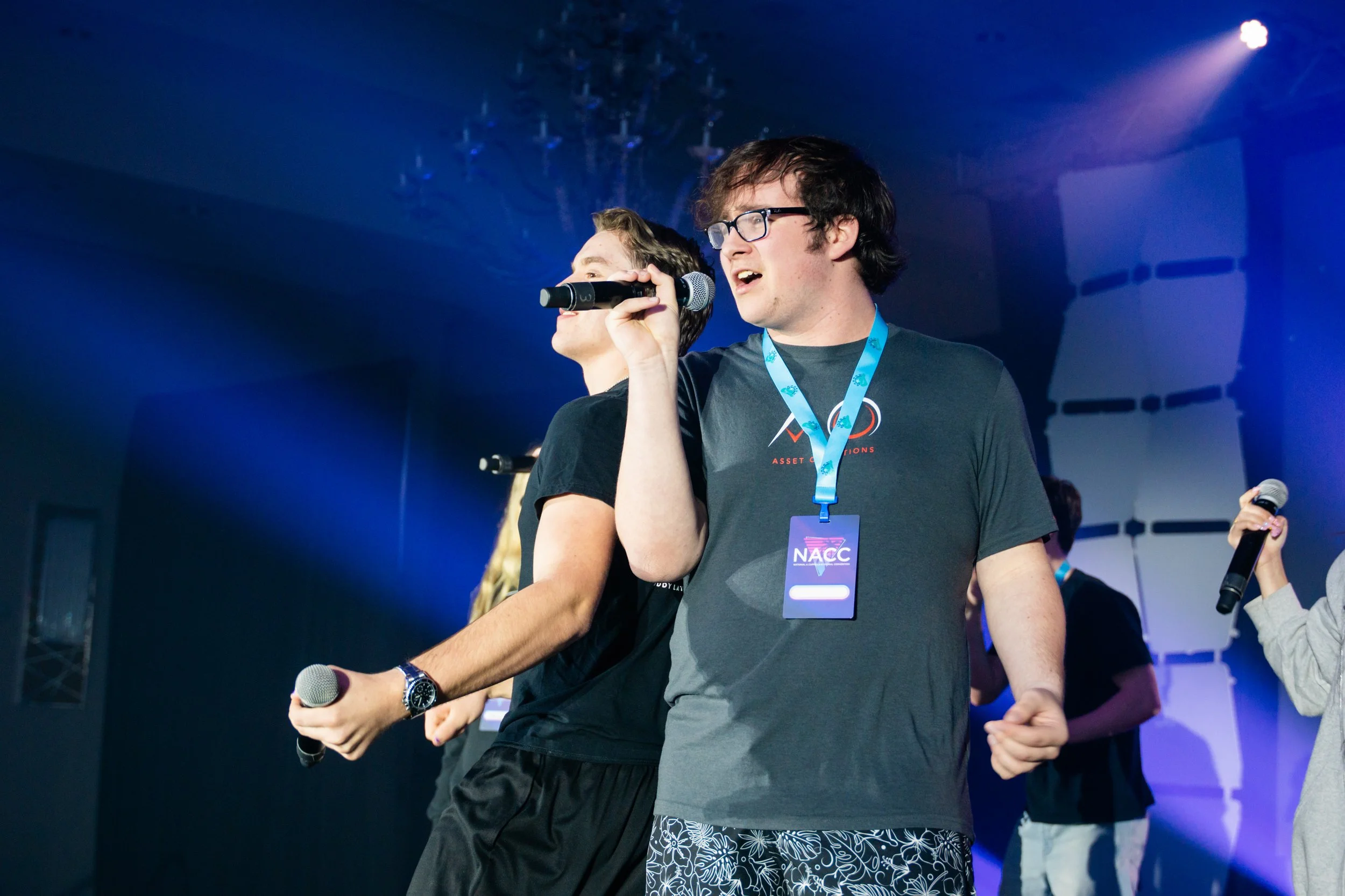 Young man with glasses singing into a microphone at a concert, wearing a lanyard with an event badge, surrounded by other people on stage.