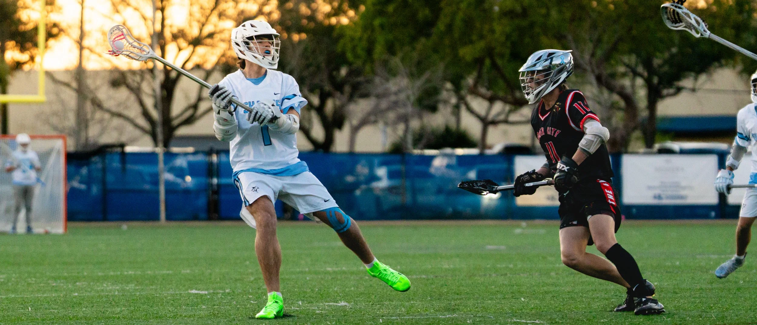 Two lacrosse players on a field, one in a white uniform and the other in a black uniform, competing for the ball during a game, with trees and a goalpost in the background.