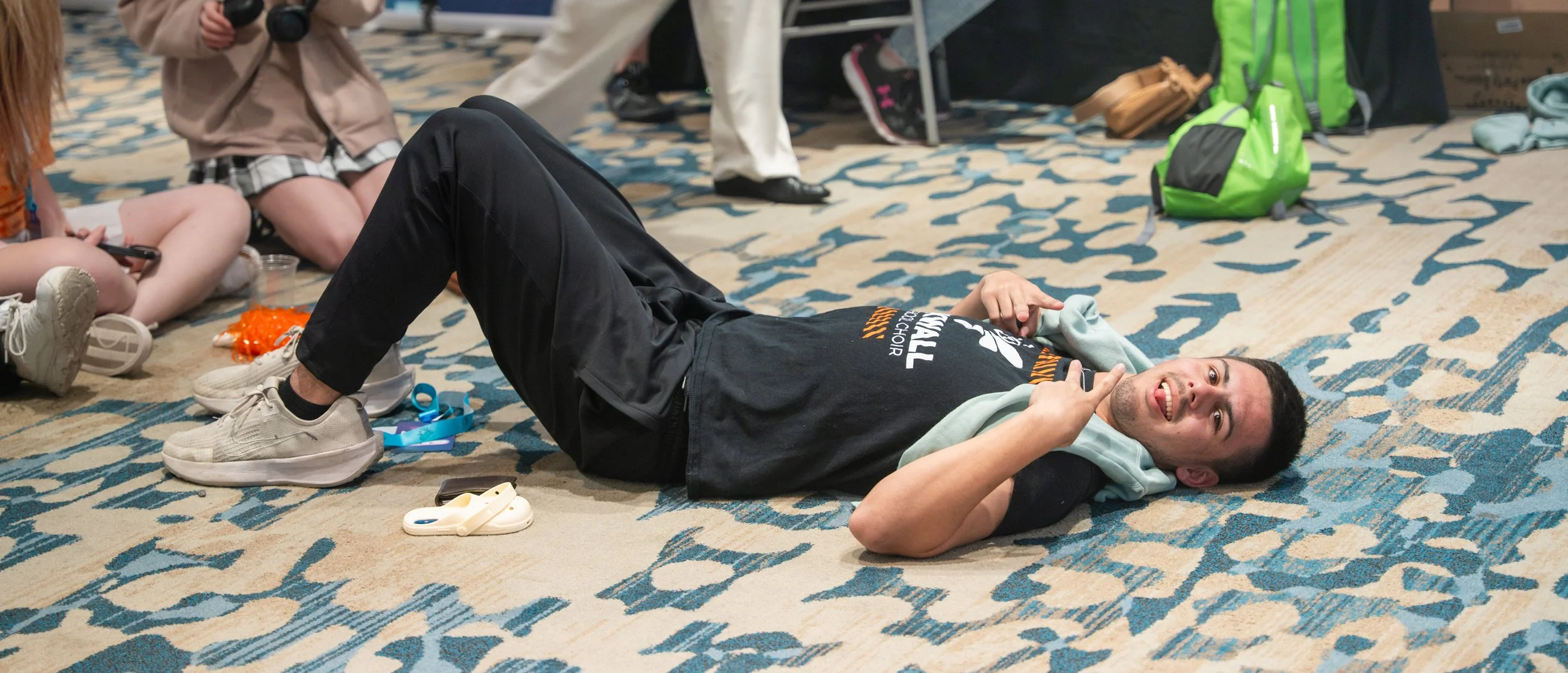 A young man lying on his side on a carpeted floor, smiling and looking at the camera, with other people sitting around him, some with cameras. Various personal items and backpacks are visible nearby.