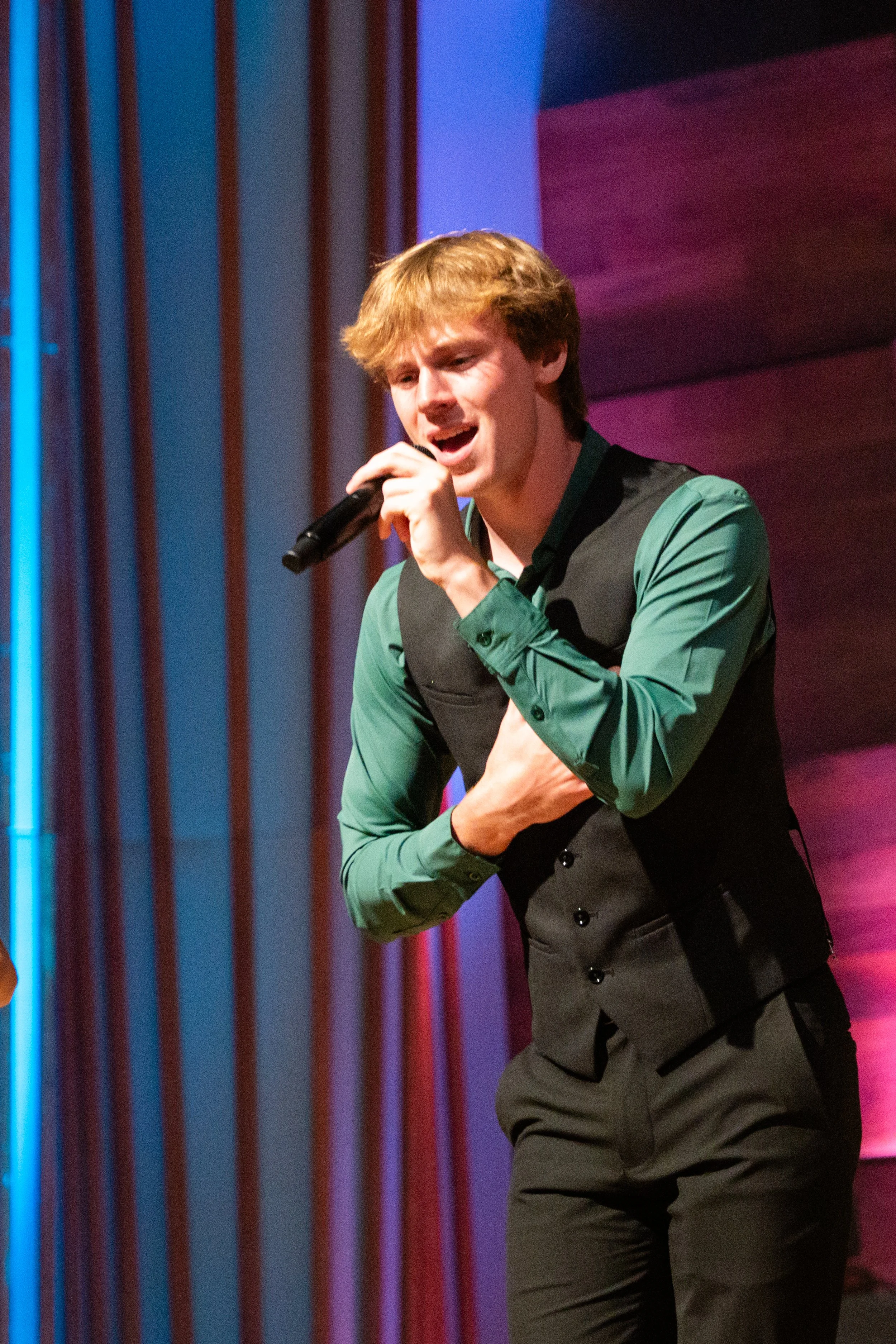 A young man singing into a microphone on stage, wearing a green shirt and black vest, with a colorful background.