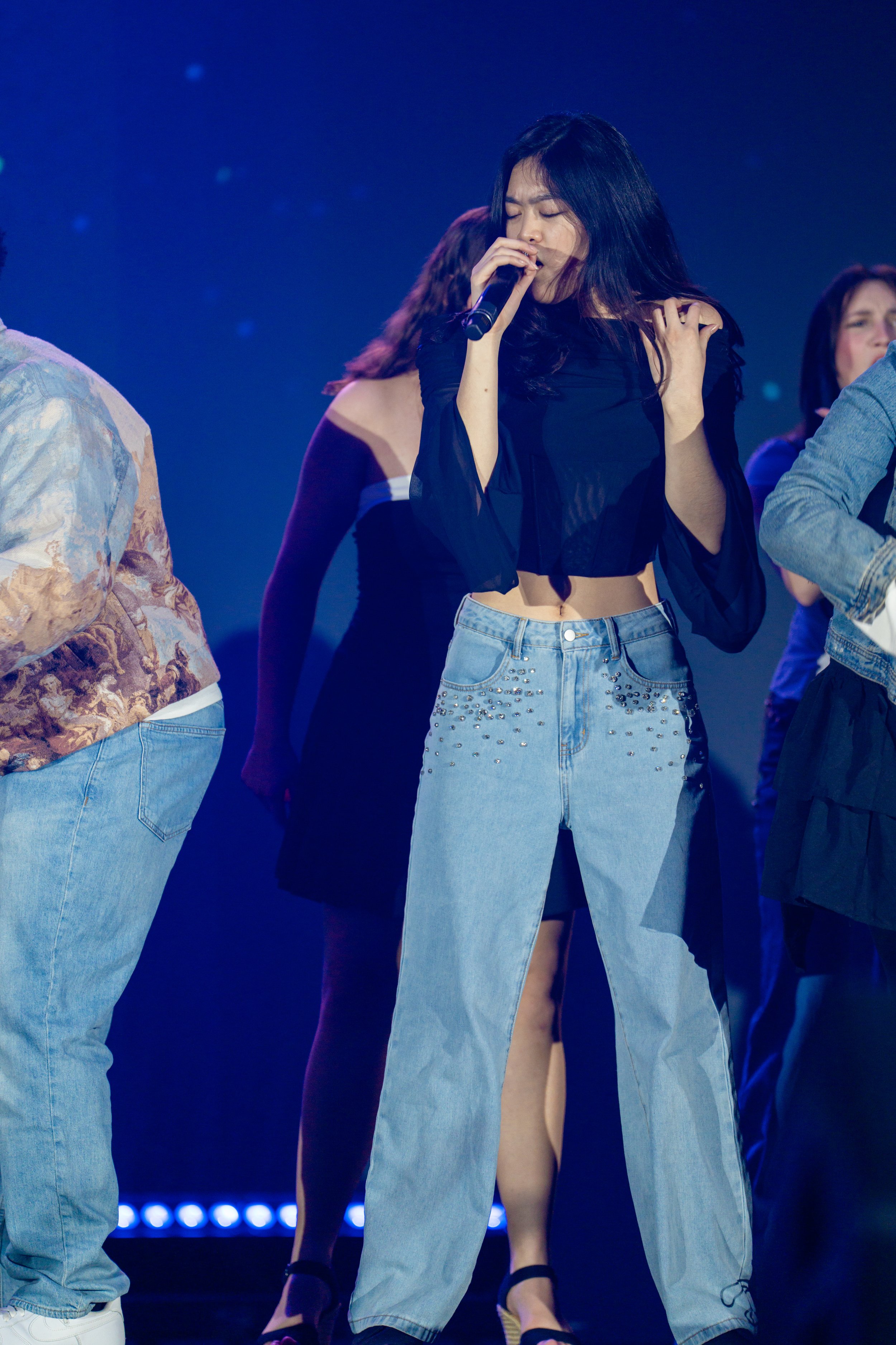 A young woman singing with her eyes closed on stage, wearing a black top and high-waisted jeans with a slit.