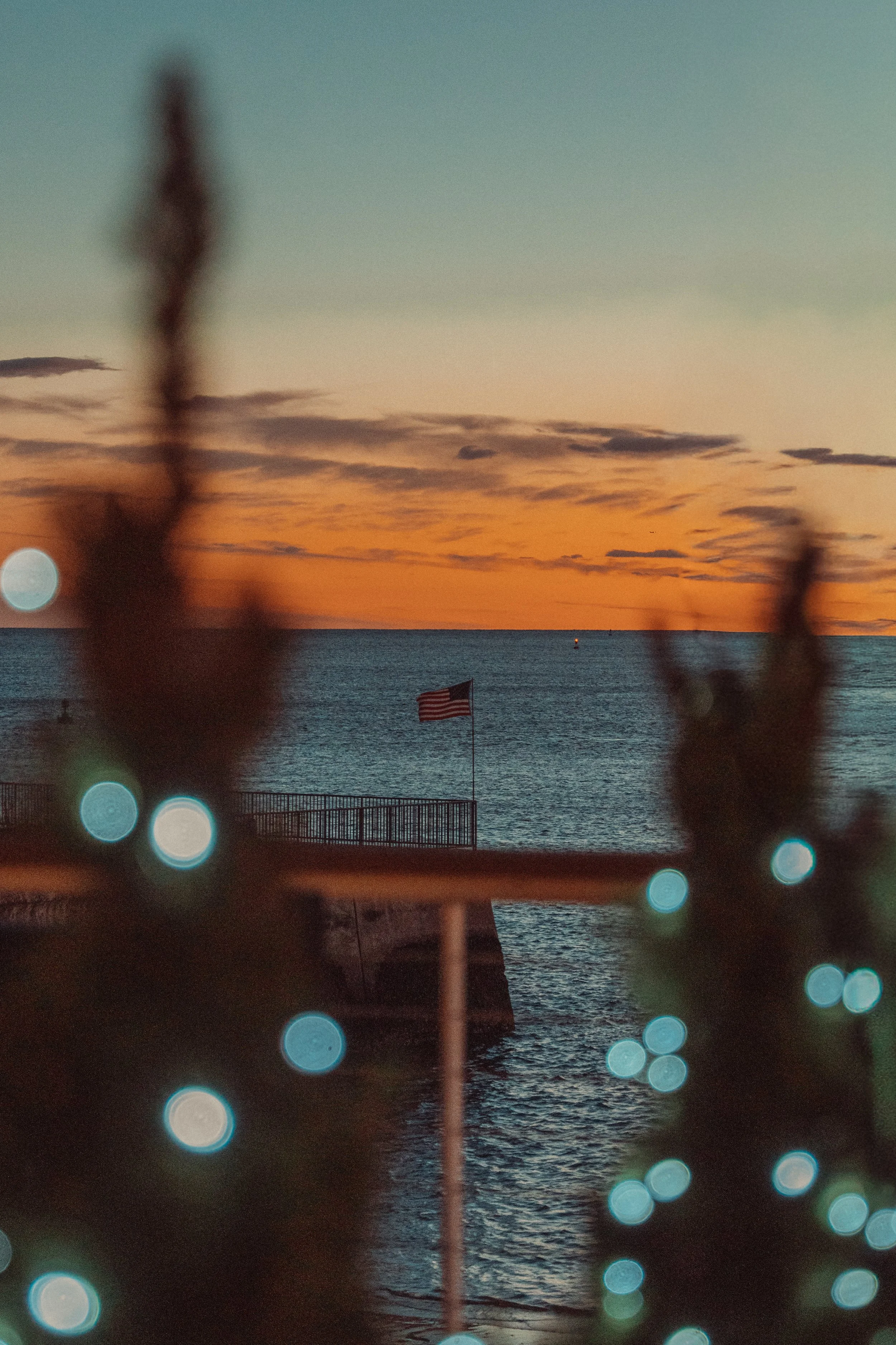 A sunset over the ocean with an American flag on a pier, blurred foreground lights, and some foliage.