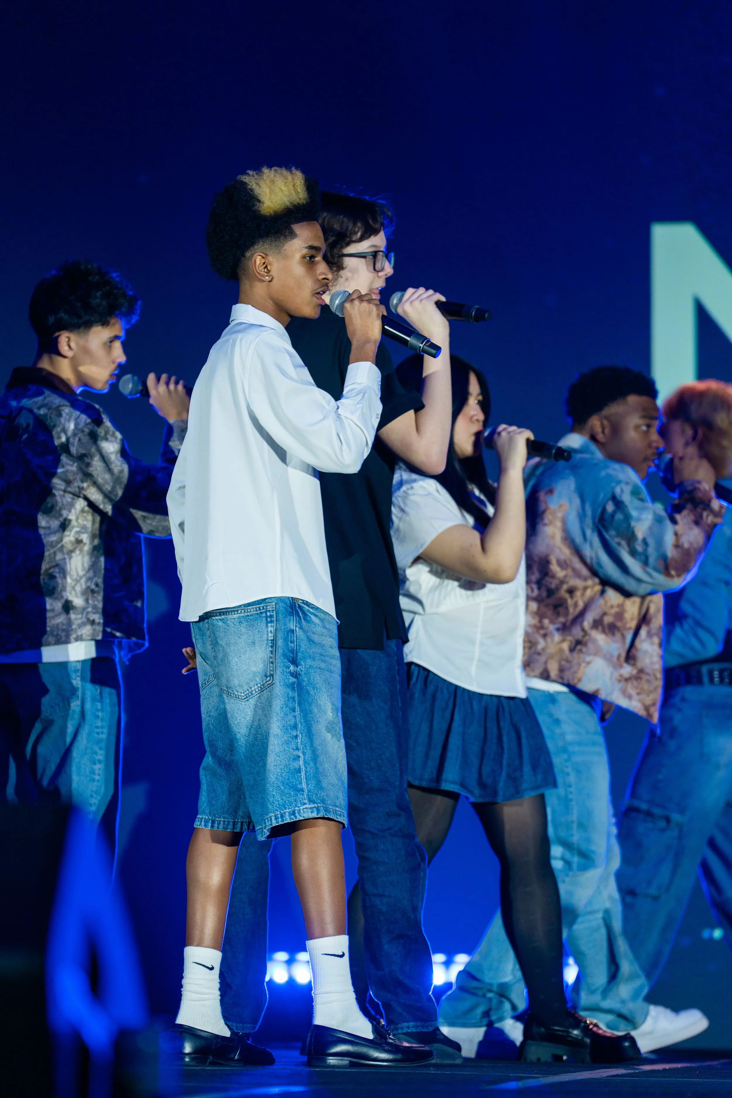 A group of young people standing on stage, holding microphones as they perform or speak. The stage is lit with blue lighting.