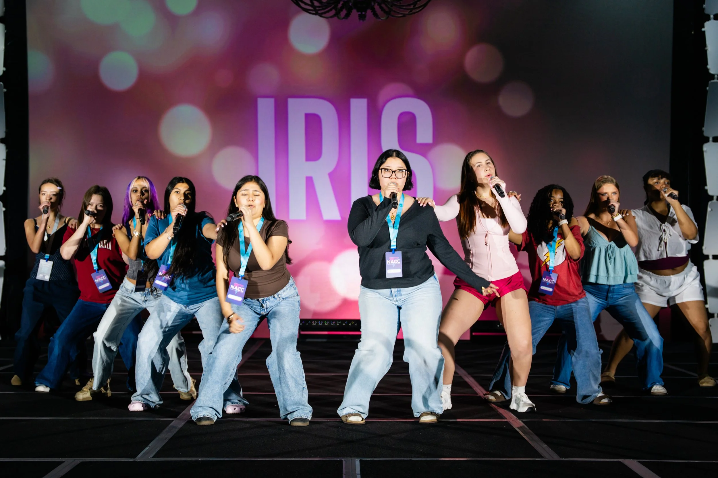 A diverse group of women performing on stage, singing into microphones with a large screen behind them displaying the text 'IDLC' against a colorful, bokeh-style background.