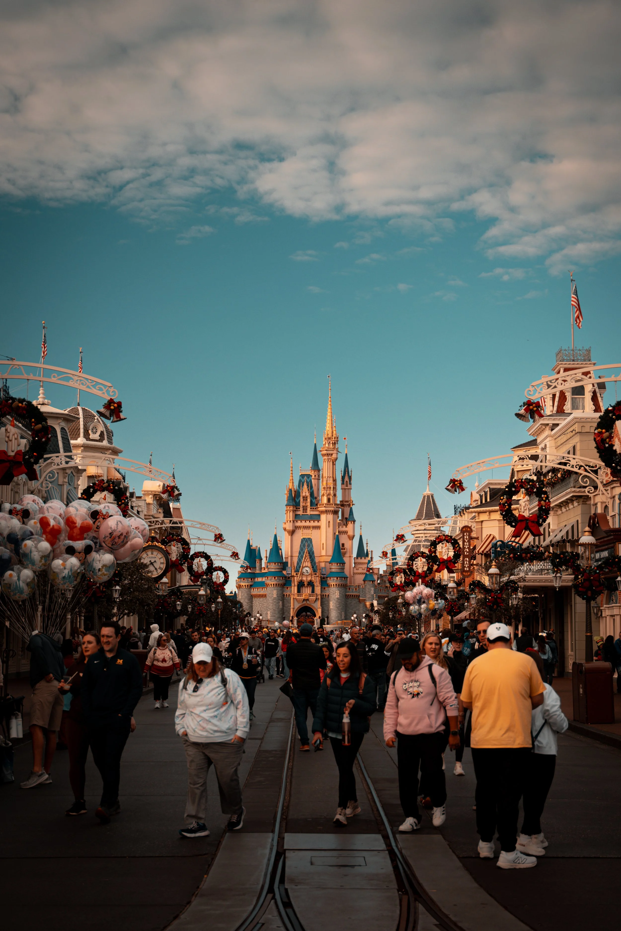 People walking on Main Street at Disneyland with Sleeping Beauty Castle in the background, decorated with holiday wreaths and balloons.