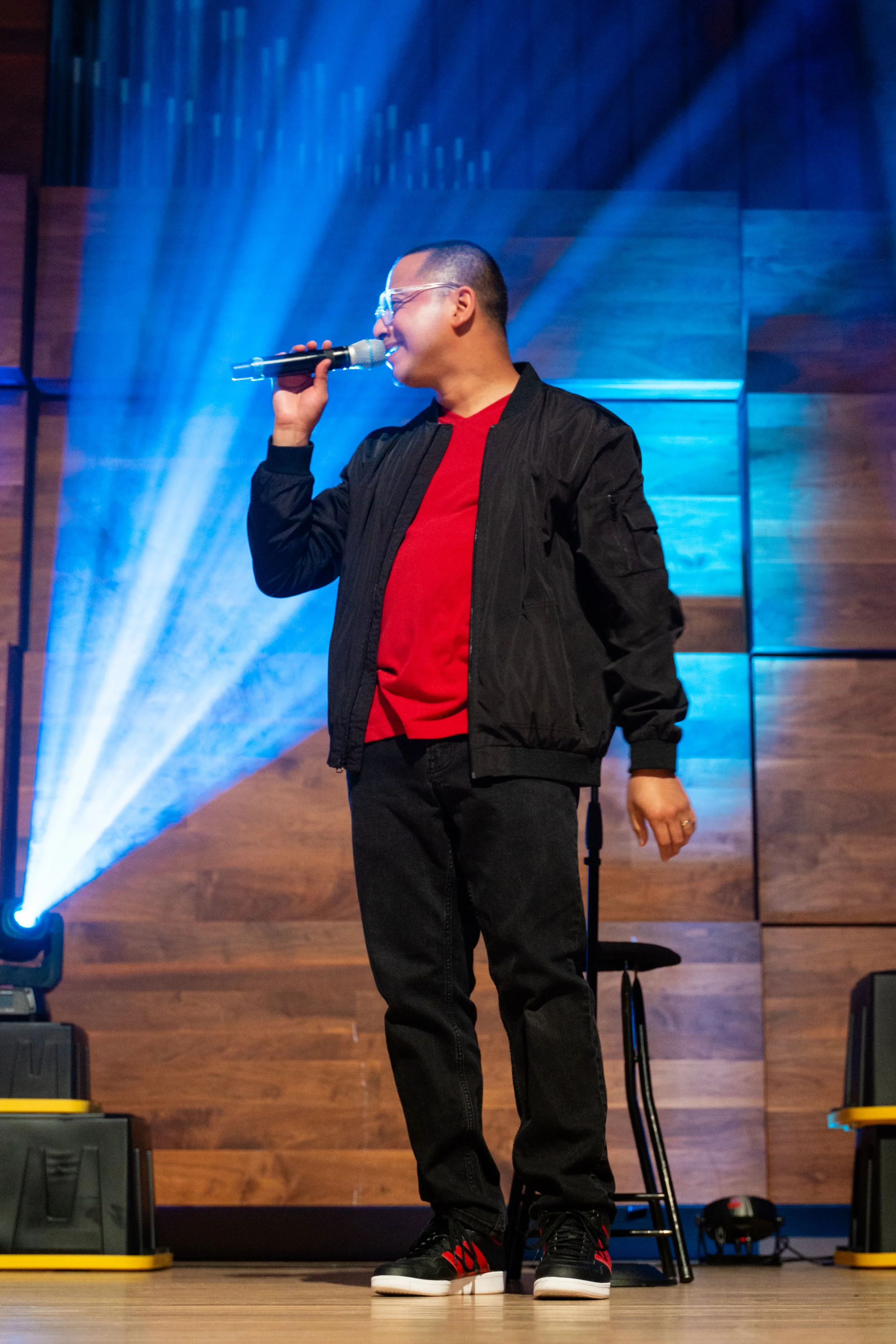 A man wearing glasses, a black jacket, and a red shirt is singing into a microphone on stage, with colorful stage lights and wooden wall background.