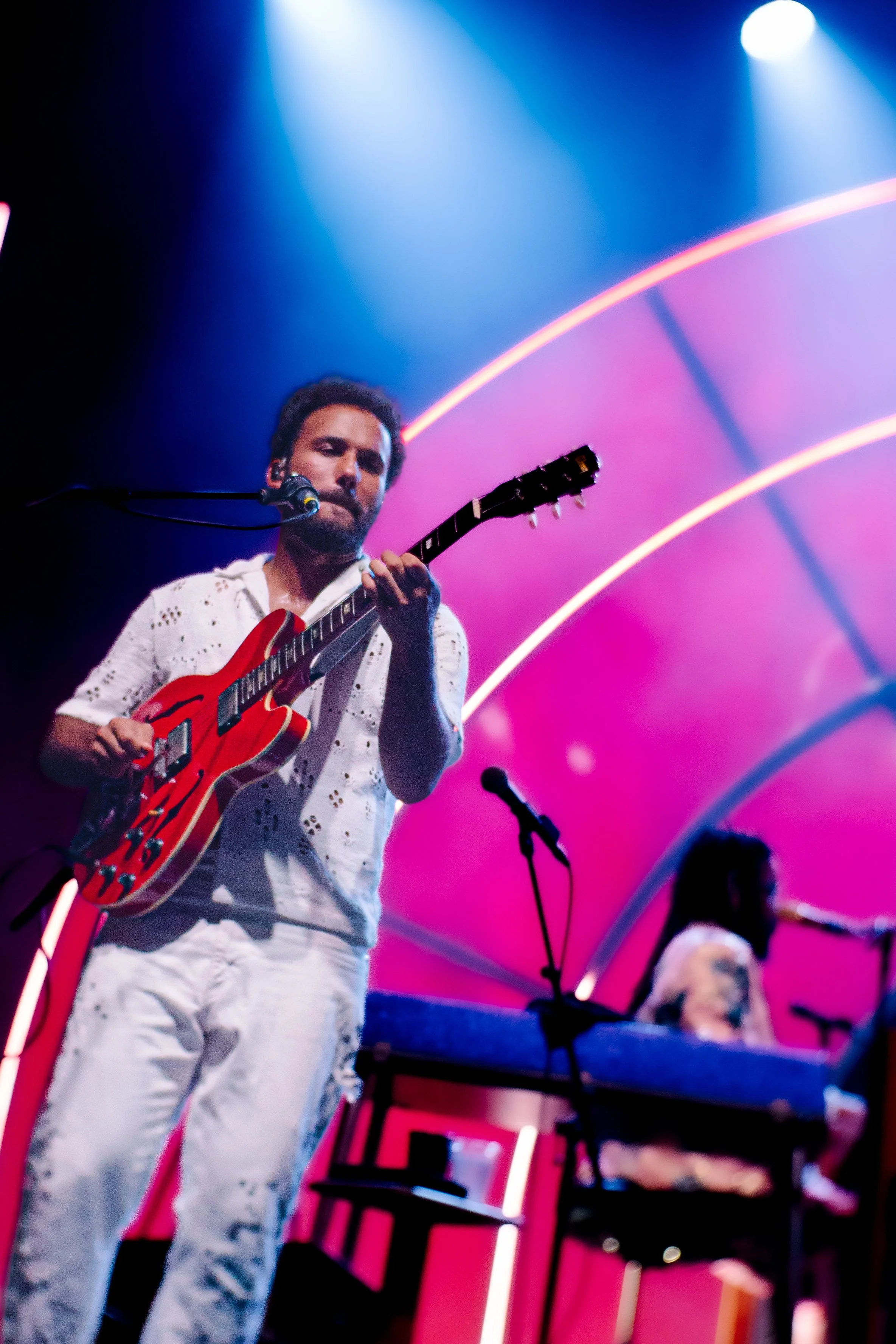 A male musician with dark hair and a beard playing an electric guitar on stage, with a female singer or musician in the background, illuminated by colorful stage lights.