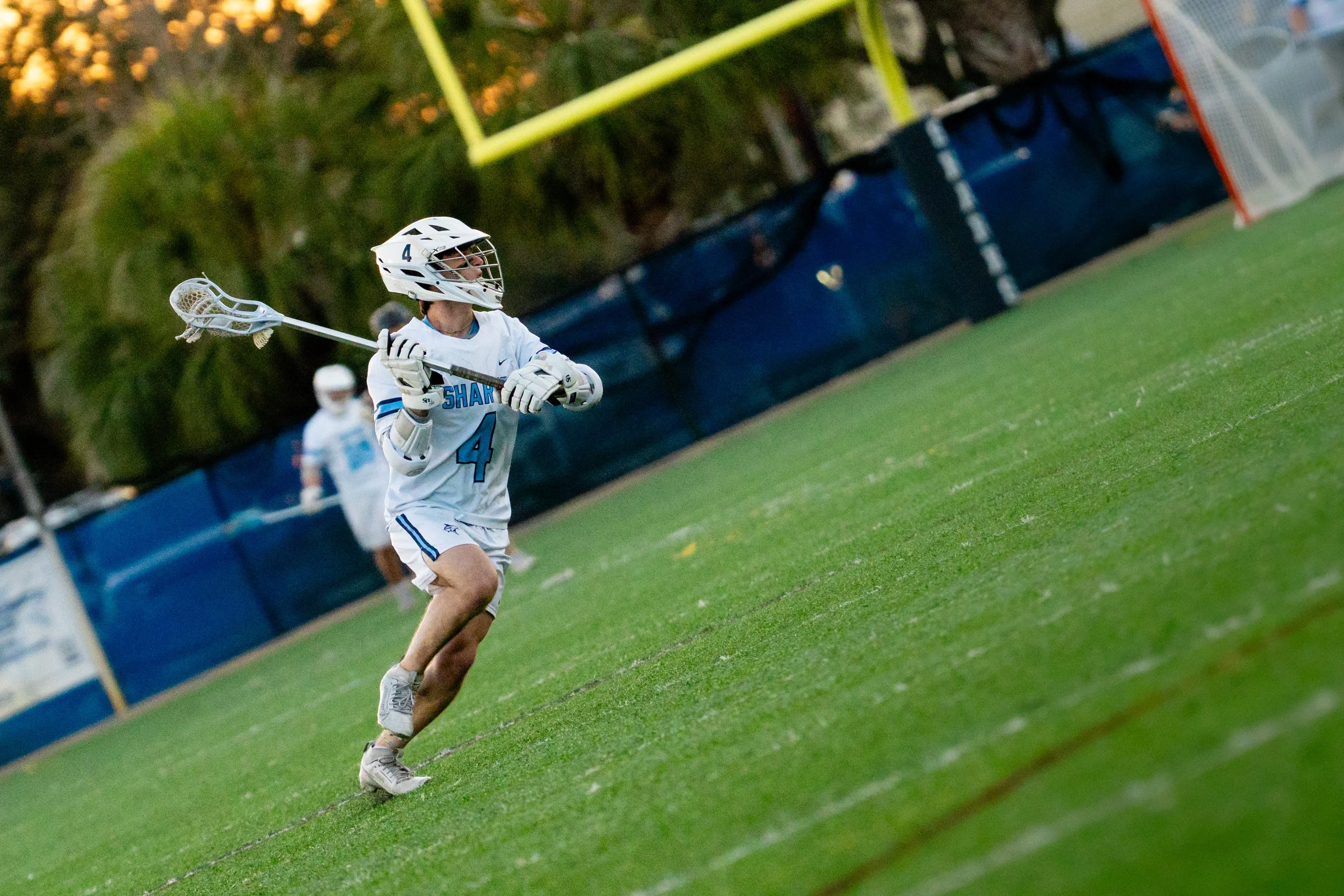 A lacrosse player wearing a white helmet with face guard, white jersey with blue accents and the number 4, is running on a green field towards the goal with a lacrosse stick in hand, in a game setting.