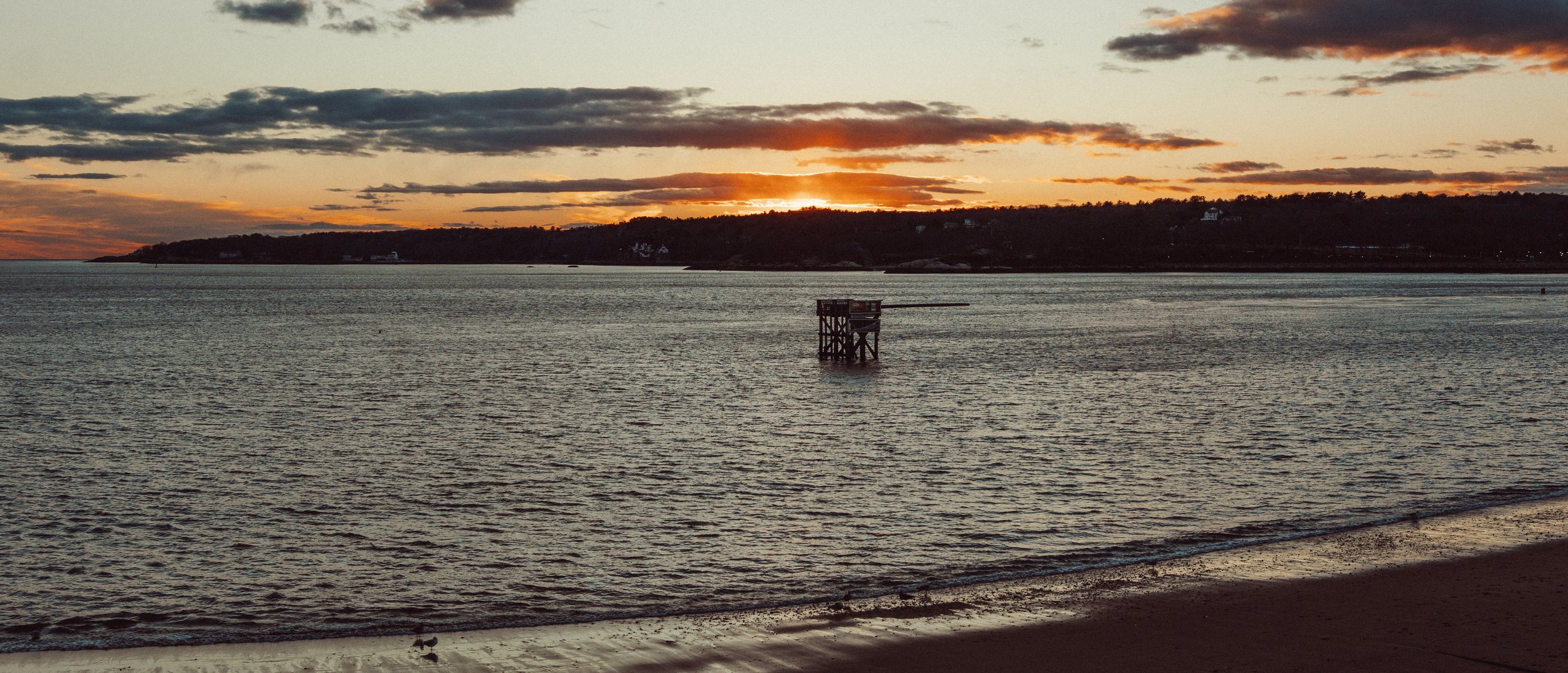 A sunset over a body of water with a distant shoreline, a small wooden structure in the water, and a sandy beach in the foreground.