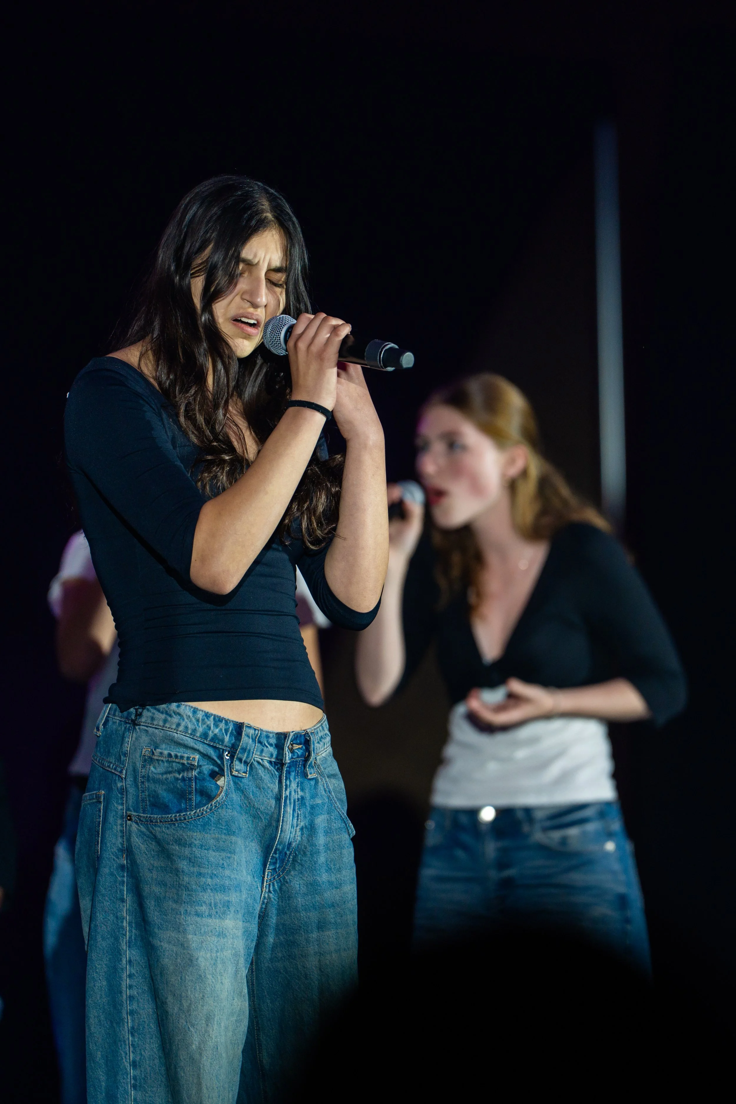 Young woman with long dark hair singing into a microphone on stage, wearing a black long-sleeve shirt and baggy jeans, with another woman in the background also singing.