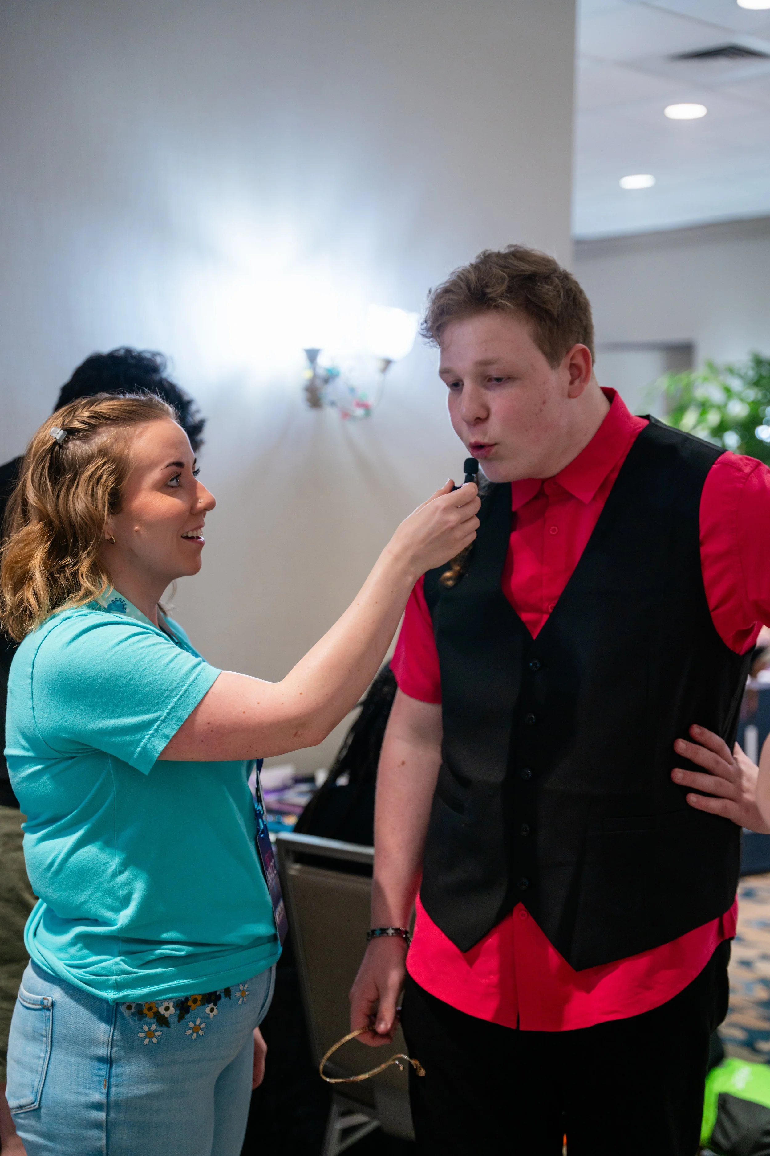 A woman with blonde hair, wearing a turquoise shirt with a daisy pattern, is holding a microphone close to a young man with red hair, wearing a red shirt and black vest. They are indoors, possibly at an event or conference, with other people and furn