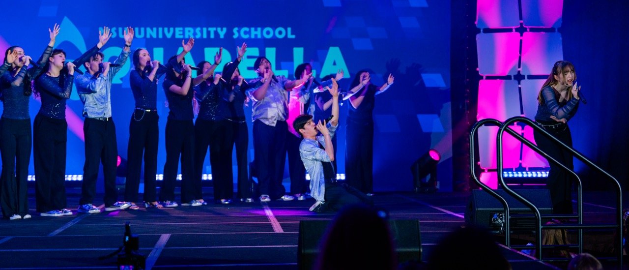Group of young performers on stage, some singing and clapping, under colorful stage lighting, with a large digital screen in the background reading "UNIVERSITY SCHOOL" and what appears to be a cheer or dance team performance.