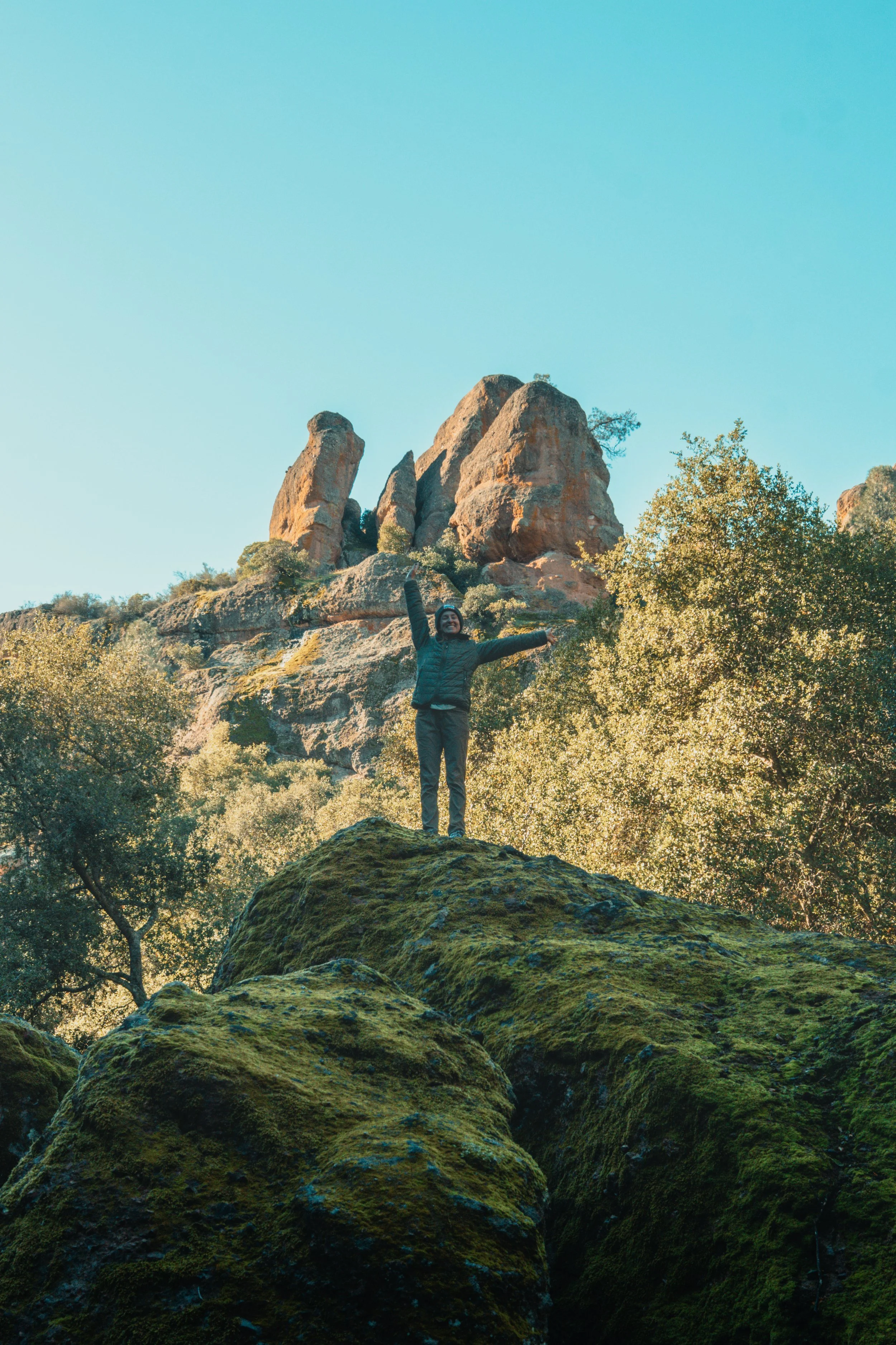 Person standing on moss-covered rocks with arms raised, surrounded by trees and large rock formations in the background under a clear blue sky.