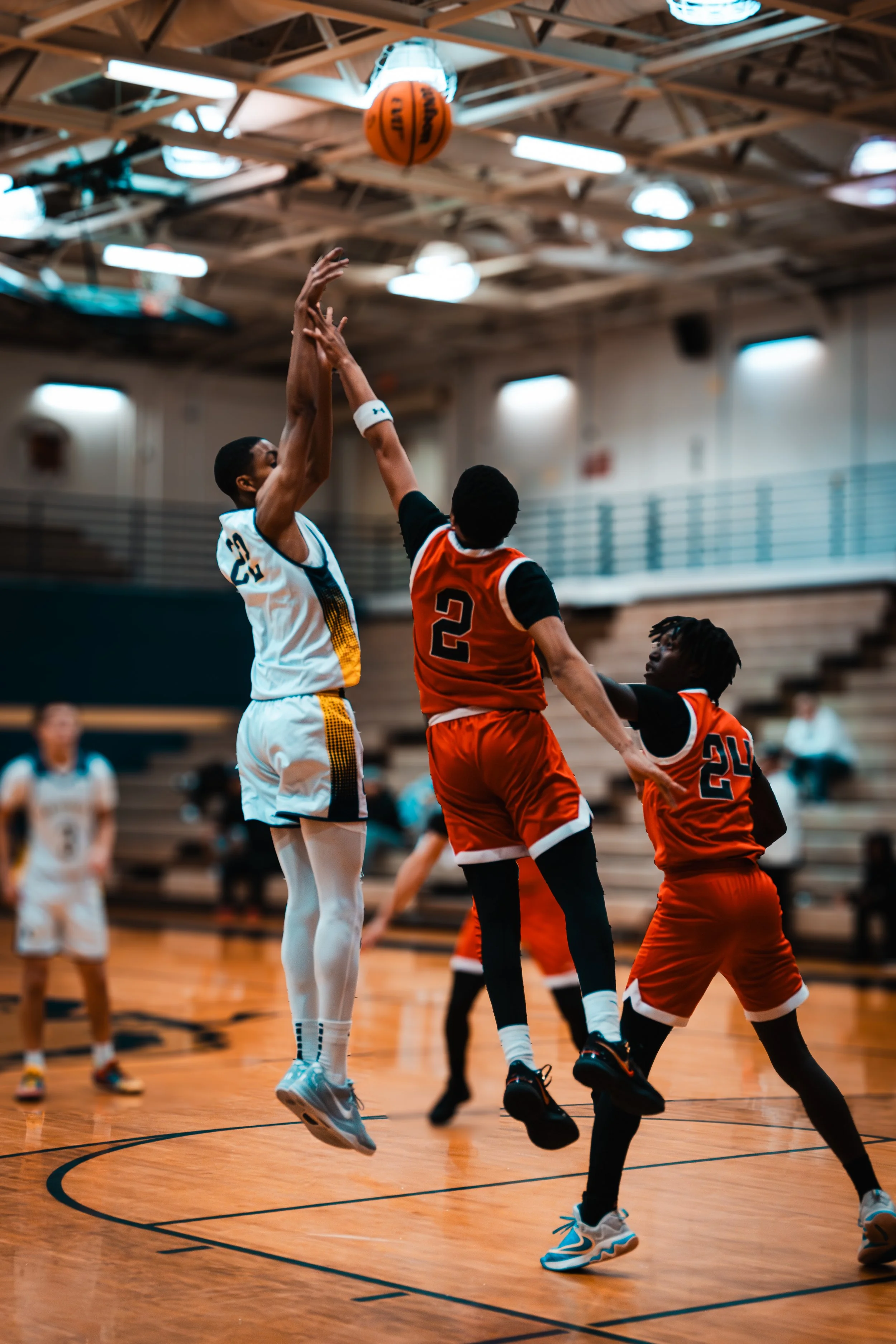 Two basketball players, one in a white uniform and one in an orange uniform, jumping to reach the basketball in an indoor gym with a wooden floor and bright ceiling lights.