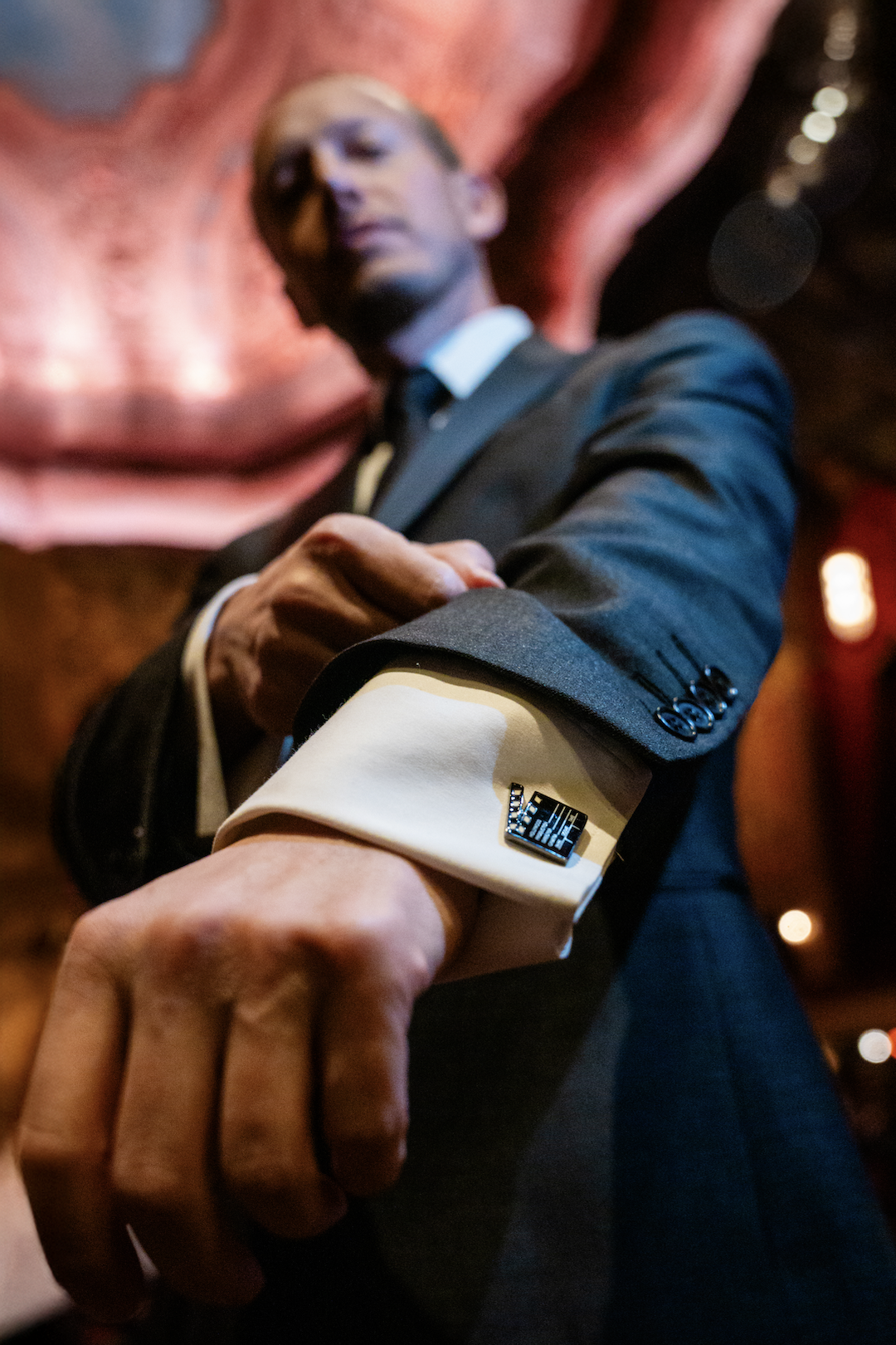 Close-up of a man in a suit adjusting his cufflink, with a low-angle perspective. The background is blurred and features warm lighting.