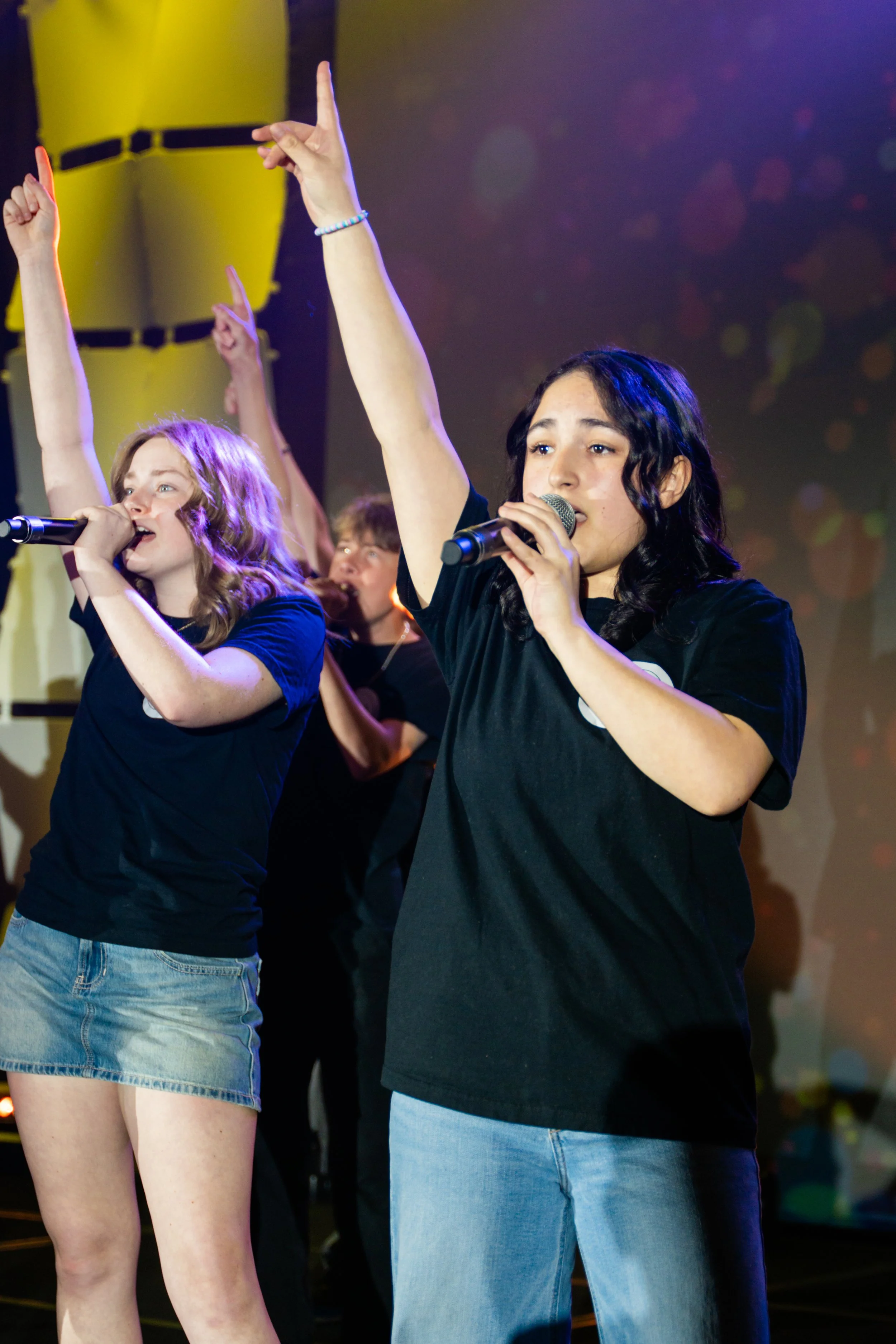 Two young women performing on stage, holding microphones, with raised hands. One has wavy red hair and wears a black t-shirt and denim shorts, while the other has curly black hair and wears a black t-shirt and blue jeans. There are additional perform