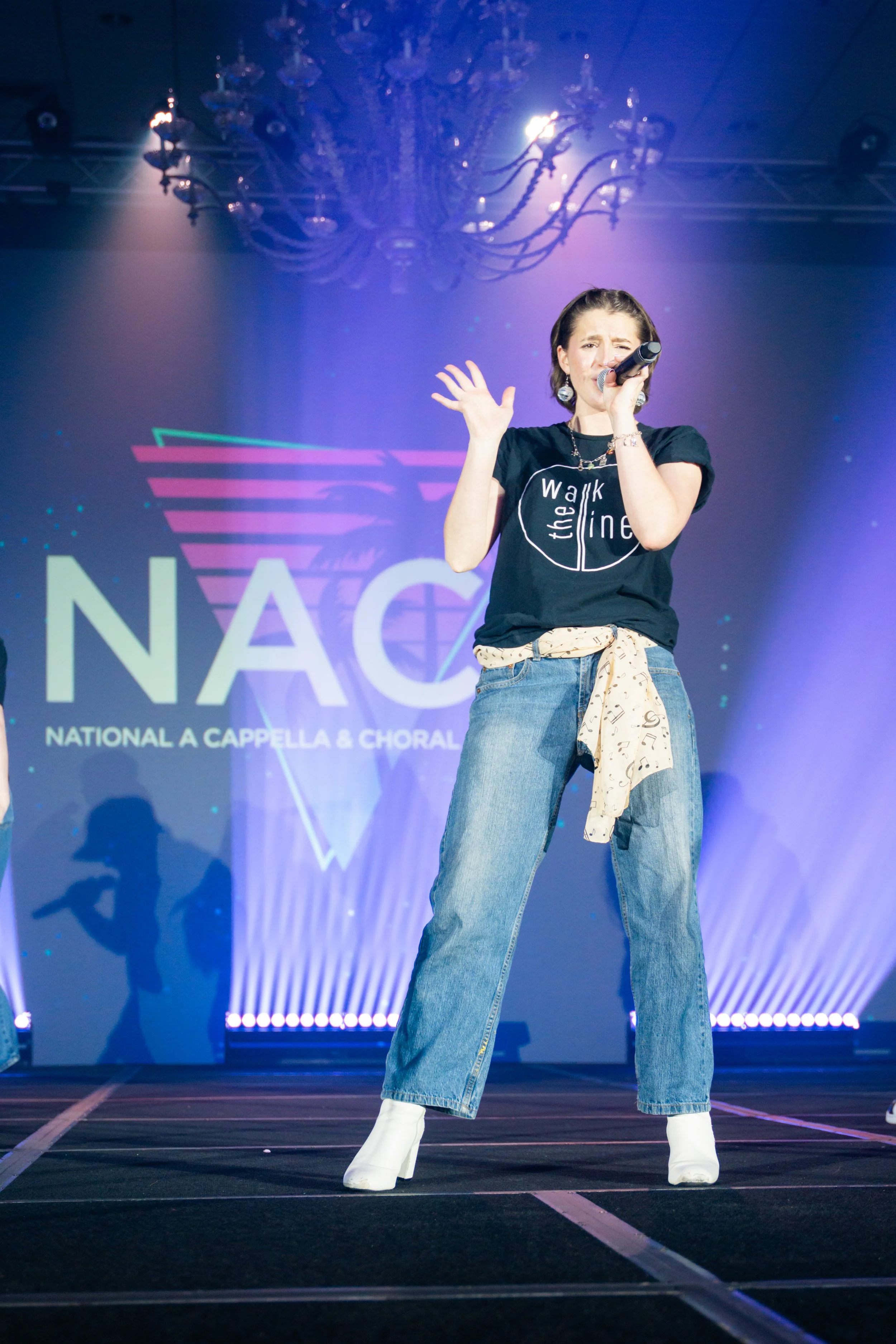 A woman singing into a microphone on stage during a performance at the National Association of Choirs event. She is wearing a black T-shirt with white text, wide-leg jeans, white boots, and has music notes tied around her waist.