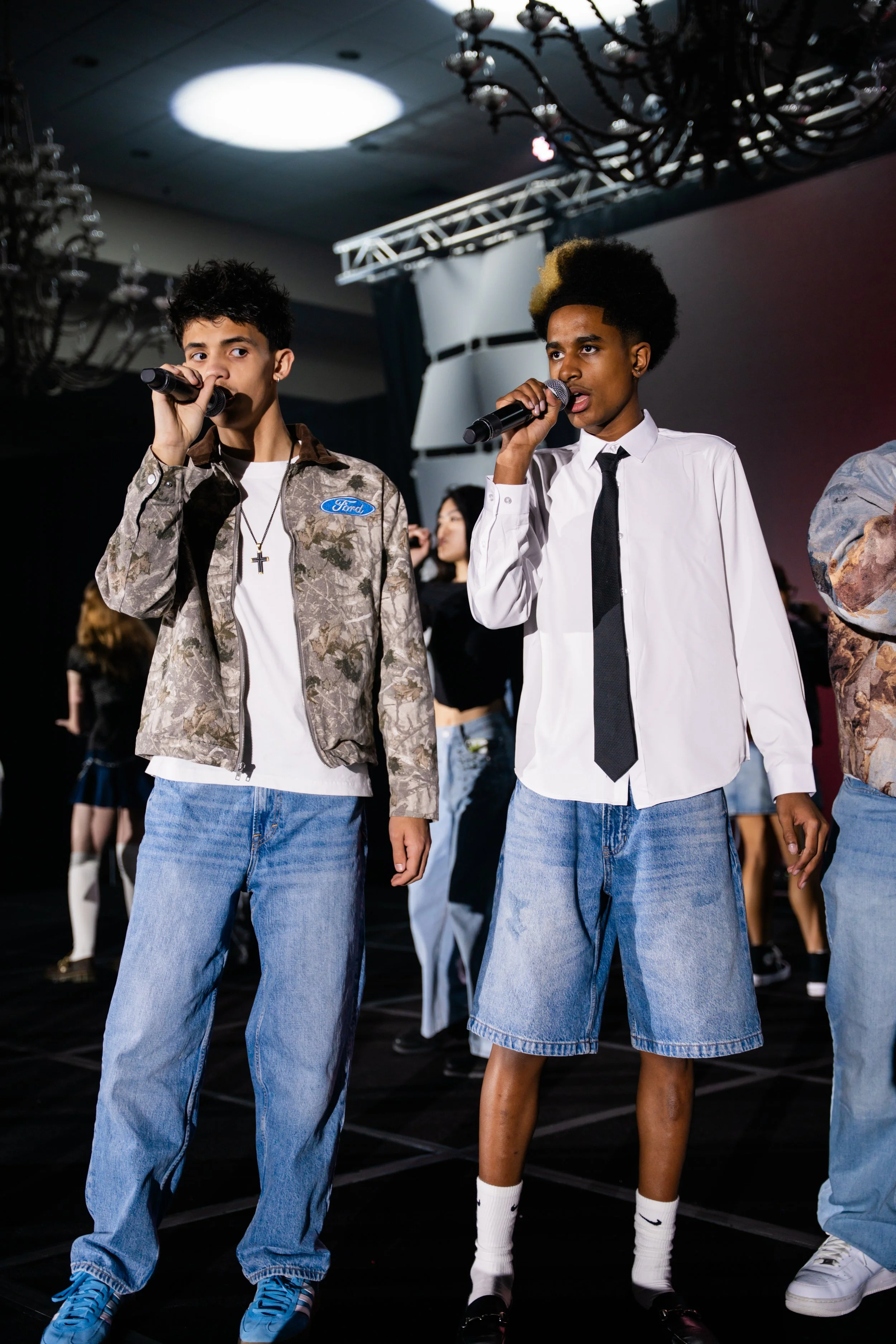 Two young women standing on a stage, each holding a microphone, with a dark background and stage lighting. One is wearing a white shirt and tie with denim shorts, and the other has a camouflage jacket with a white shirt and jeans. Multiple people are