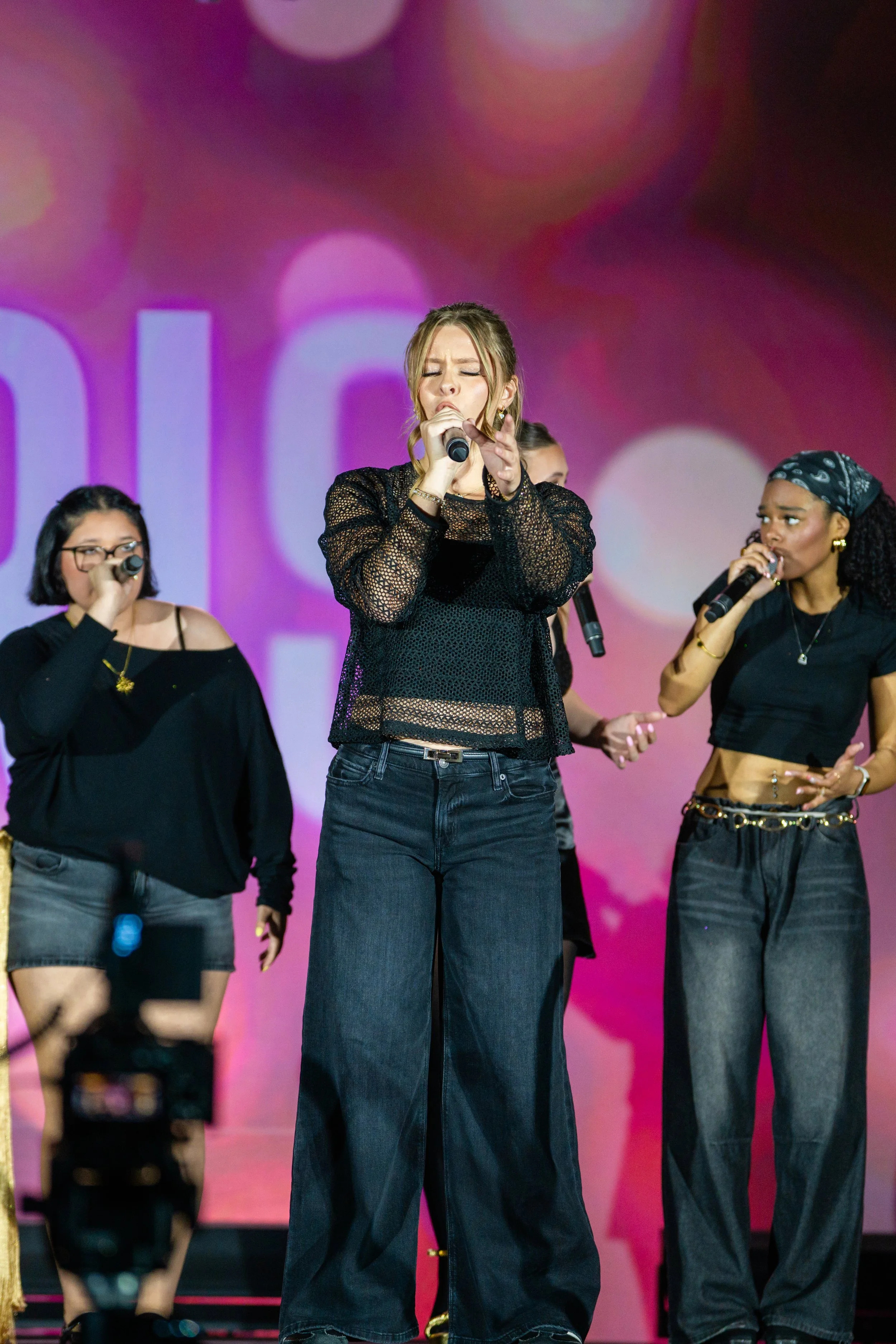 A group of women singing on stage, center woman with eyes closed, holding microphone, wearing black lace top and wide-leg jeans. Others hold microphones, dressed in dark clothing, with a pink and purple background.