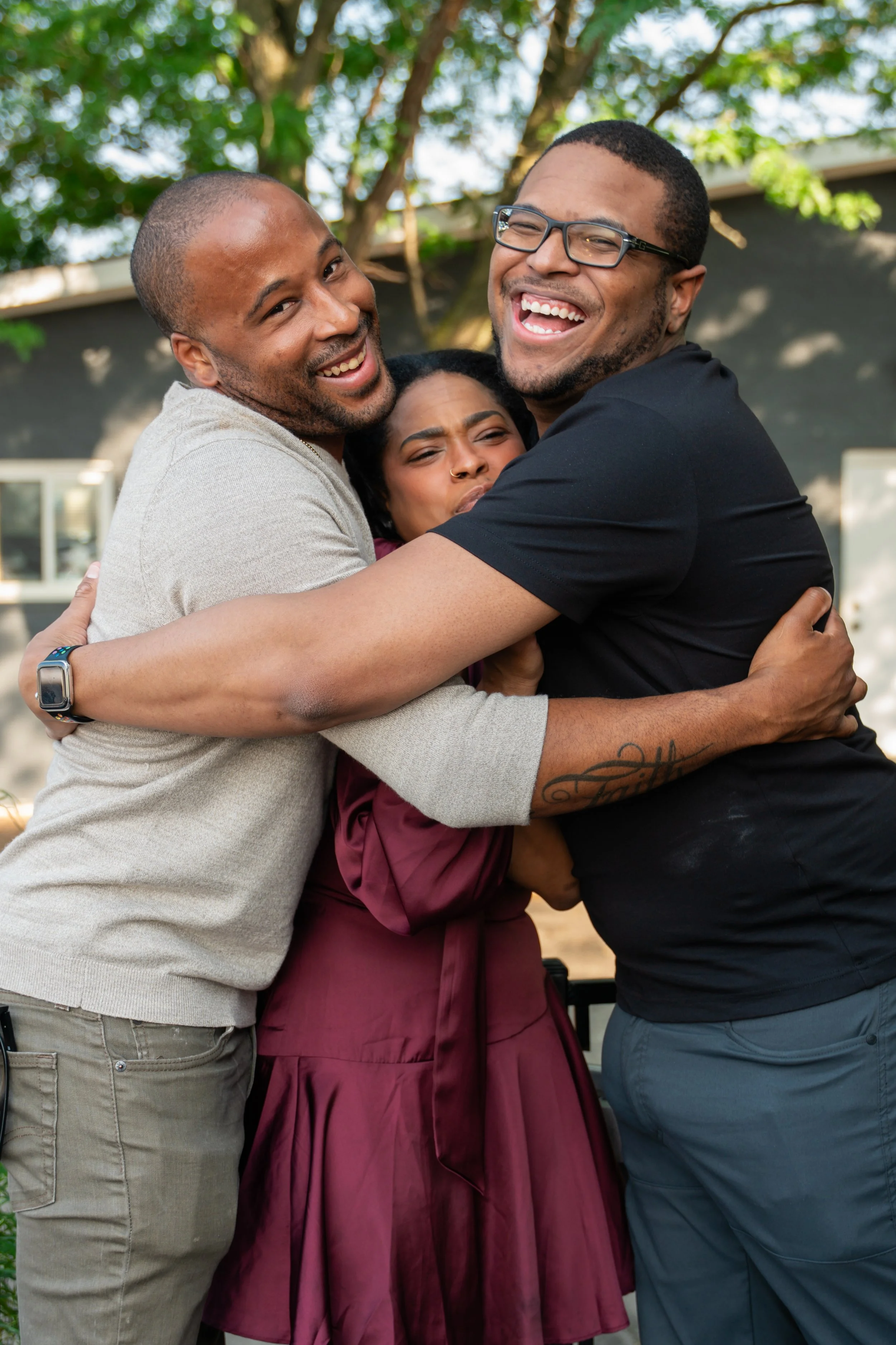 Three people are hugging and smiling outdoors, with trees and a building in the background.