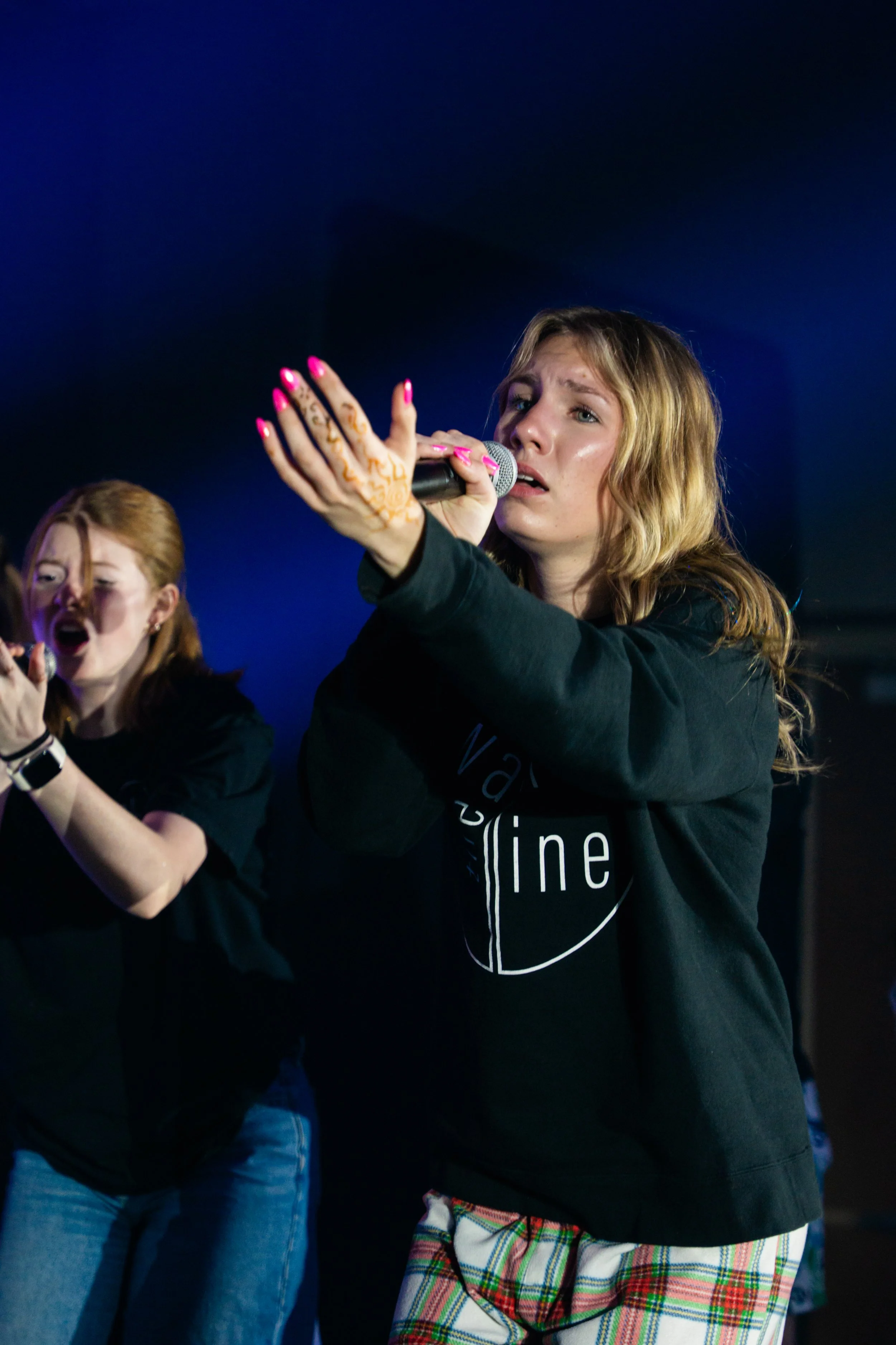 Two women singing into microphones on stage, with one wearing a black sweatshirt with writing, plaid shorts, and the other with red hair and a black shirt. The background is dark with blue lighting.