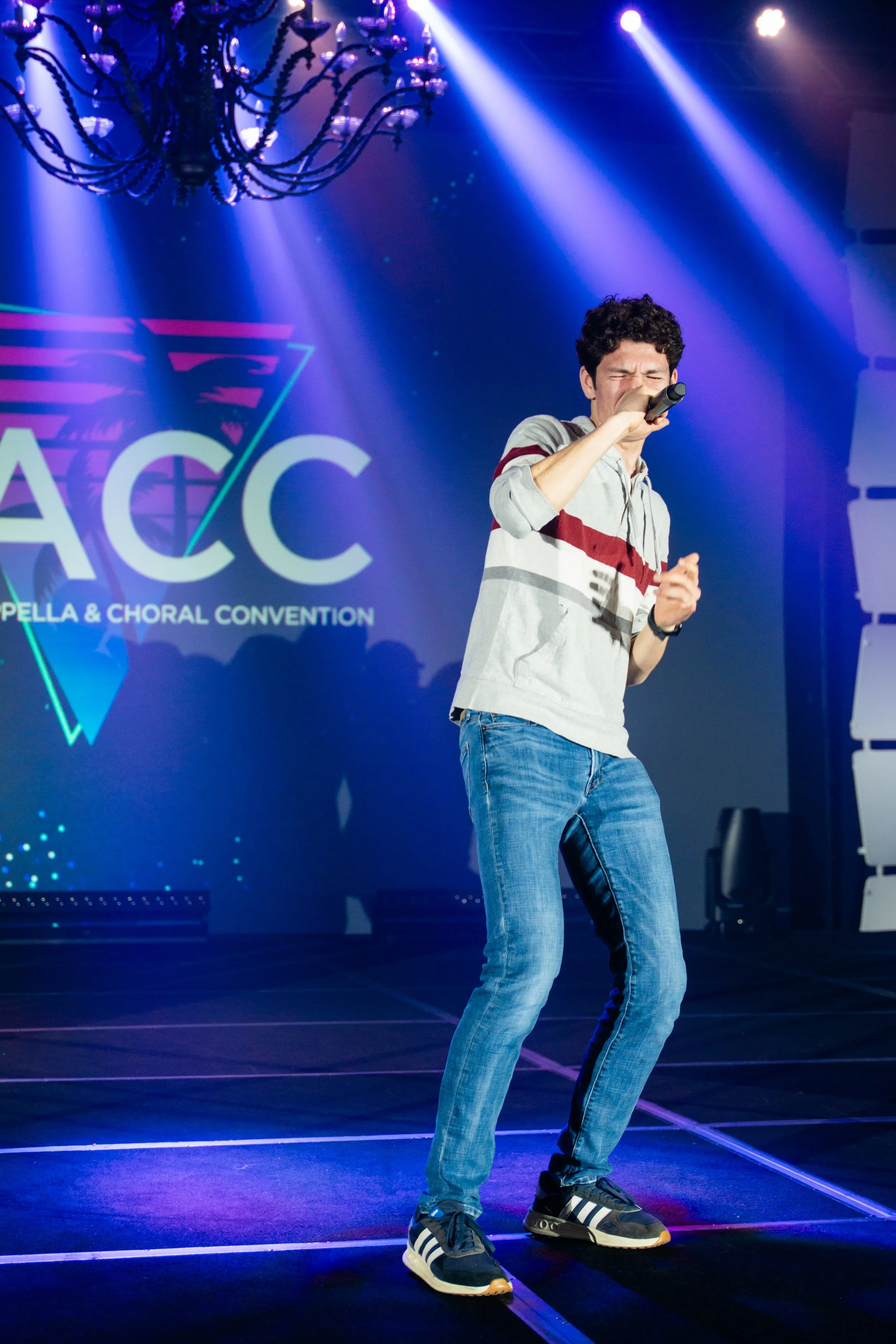 A young man singing into a microphone on stage, wearing a gray hoodie with red and black stripes and blue jeans, under blue and purple stage lights at an event called ACCA Spella & Choral Convention.