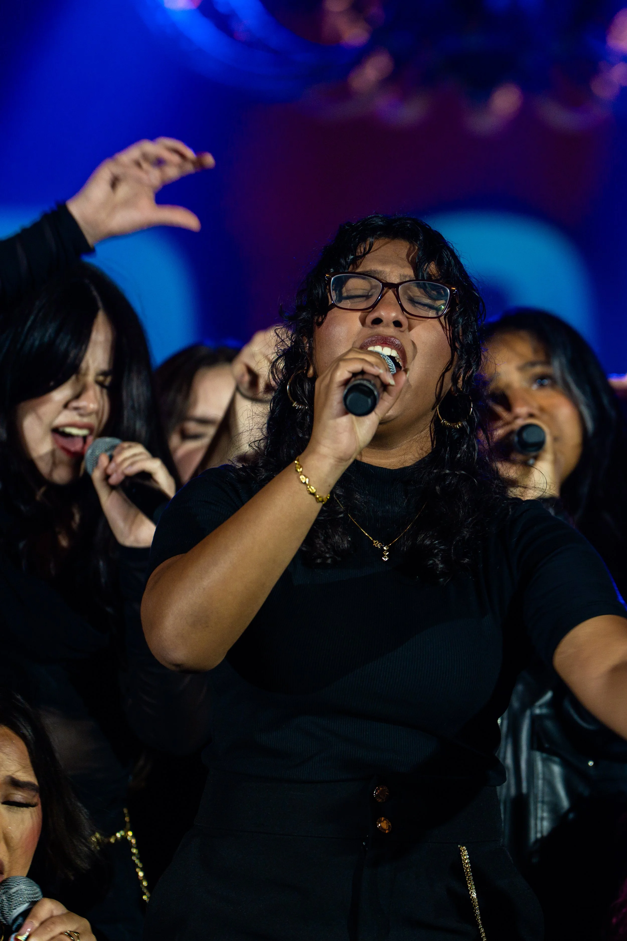 A group of women singing passionately with microphones on stage, one woman in the center wearing glasses and a black top, with blue and purple stage lighting in the background.