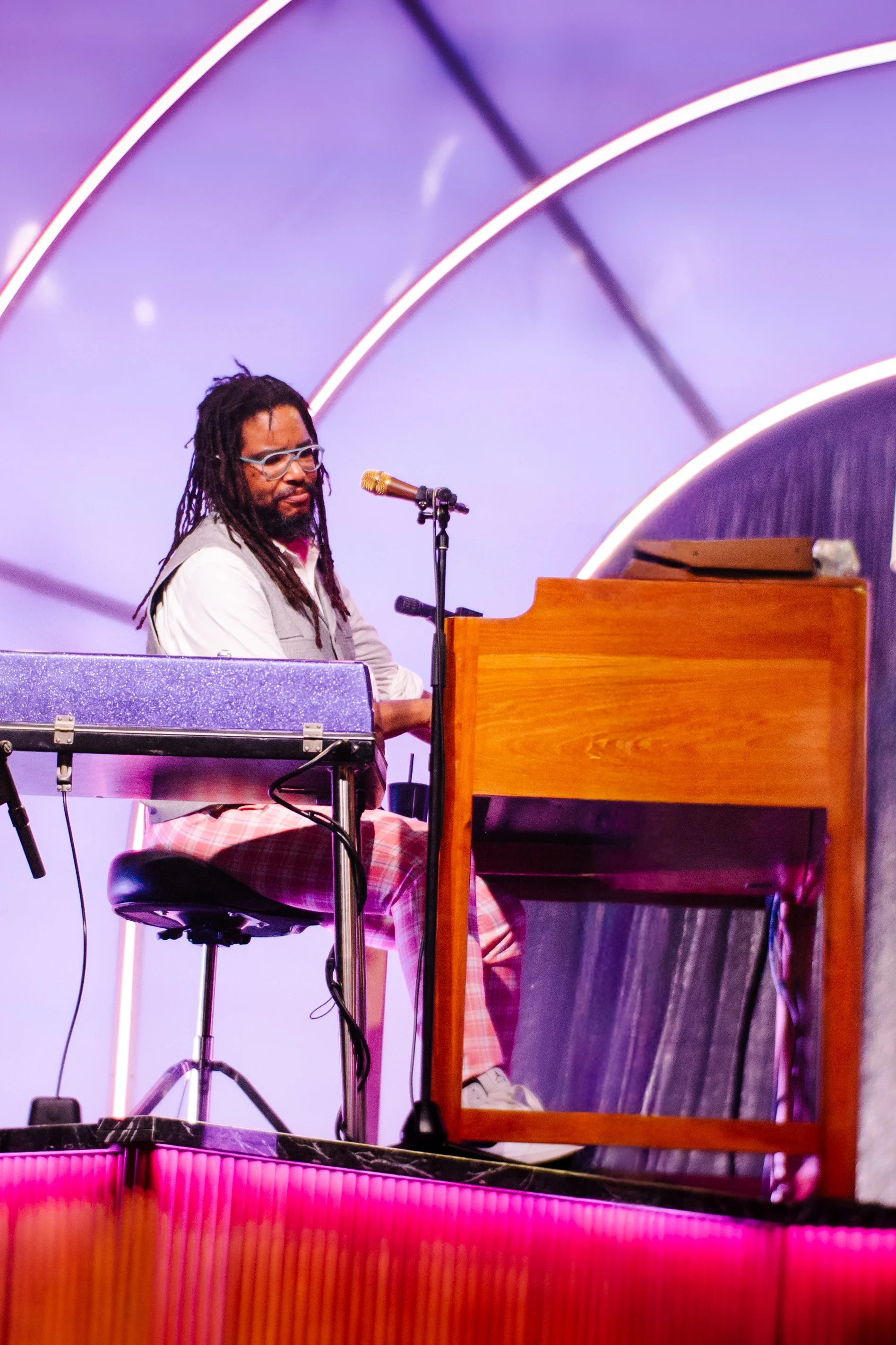Musician with dreadlocks and glasses playing piano on stage with purple and pink lighting.