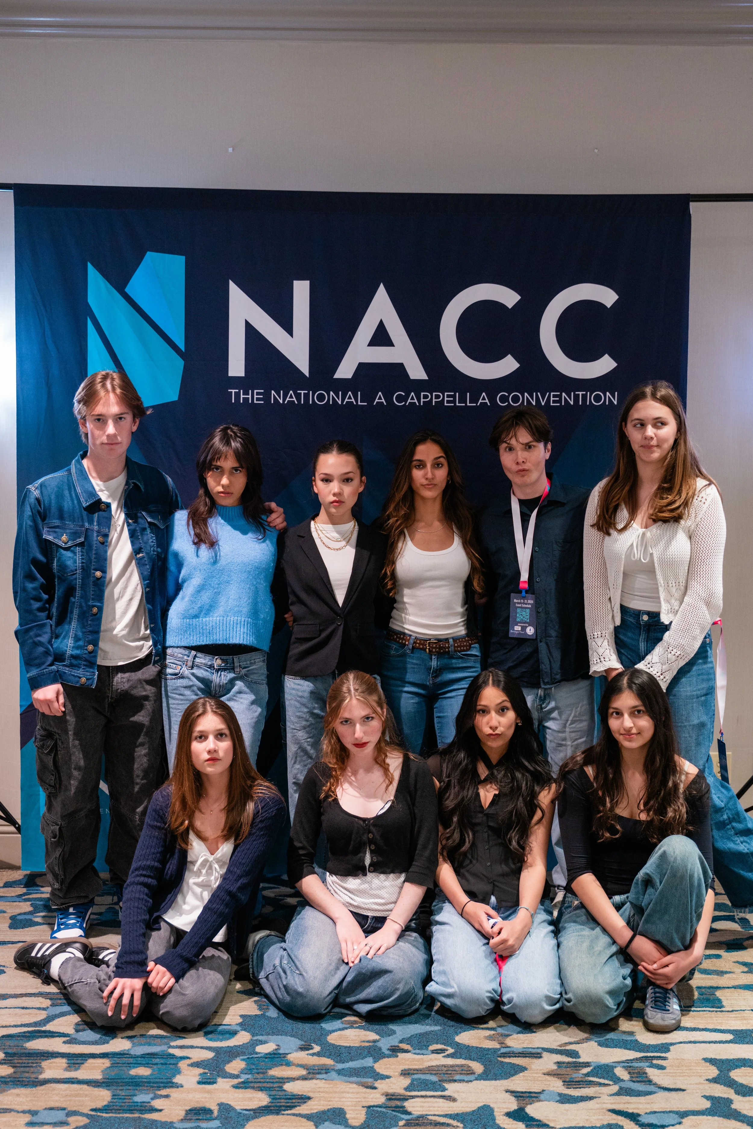 Group of young individuals posing for a photo at the National A Cappella Convention with a NAC logo banner in the background.