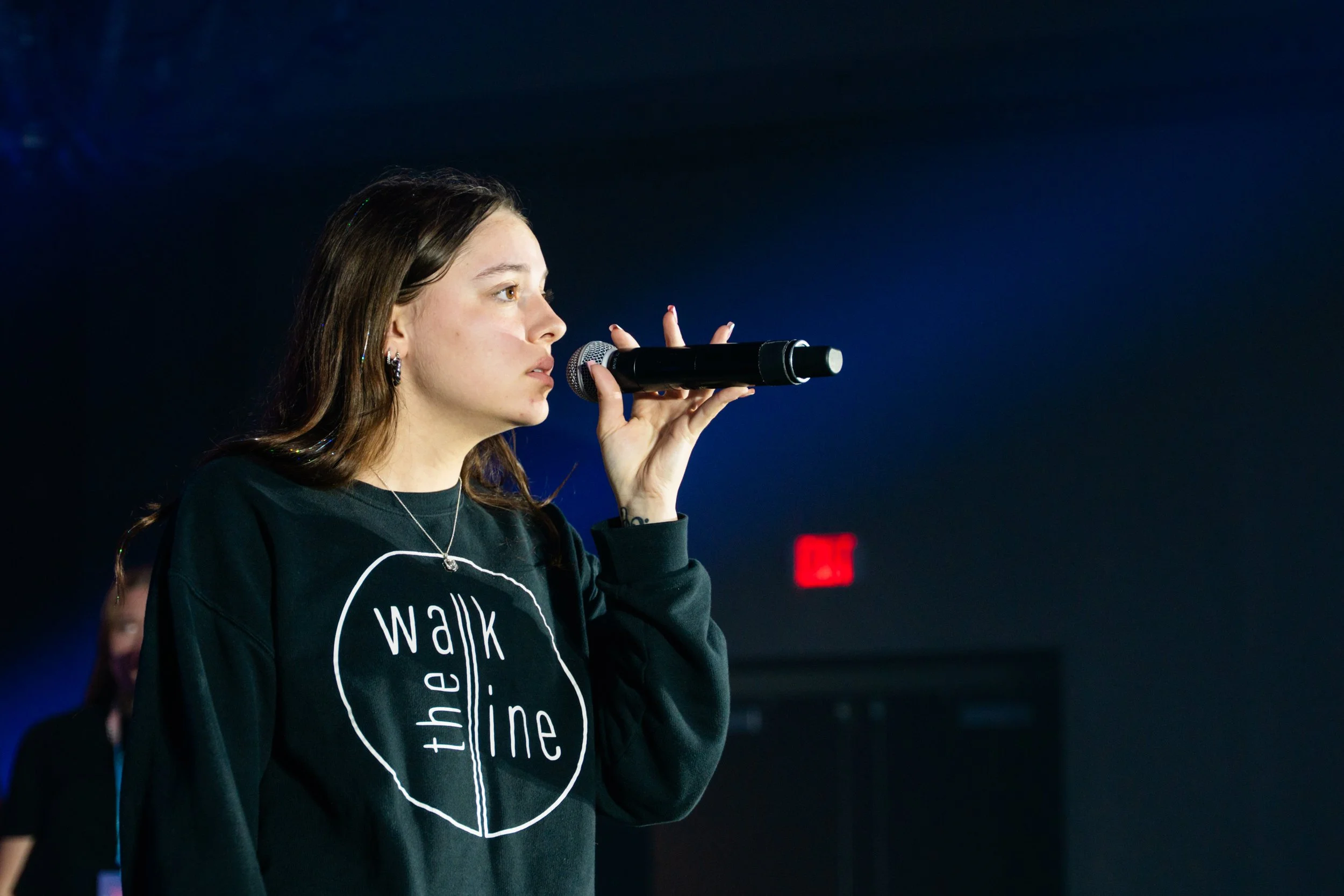 A young woman with brown hair, earrings, and a black sweatshirt holding a microphone close to her mouth, possibly speaking or singing, standing in front of a dark background with a red exit sign.