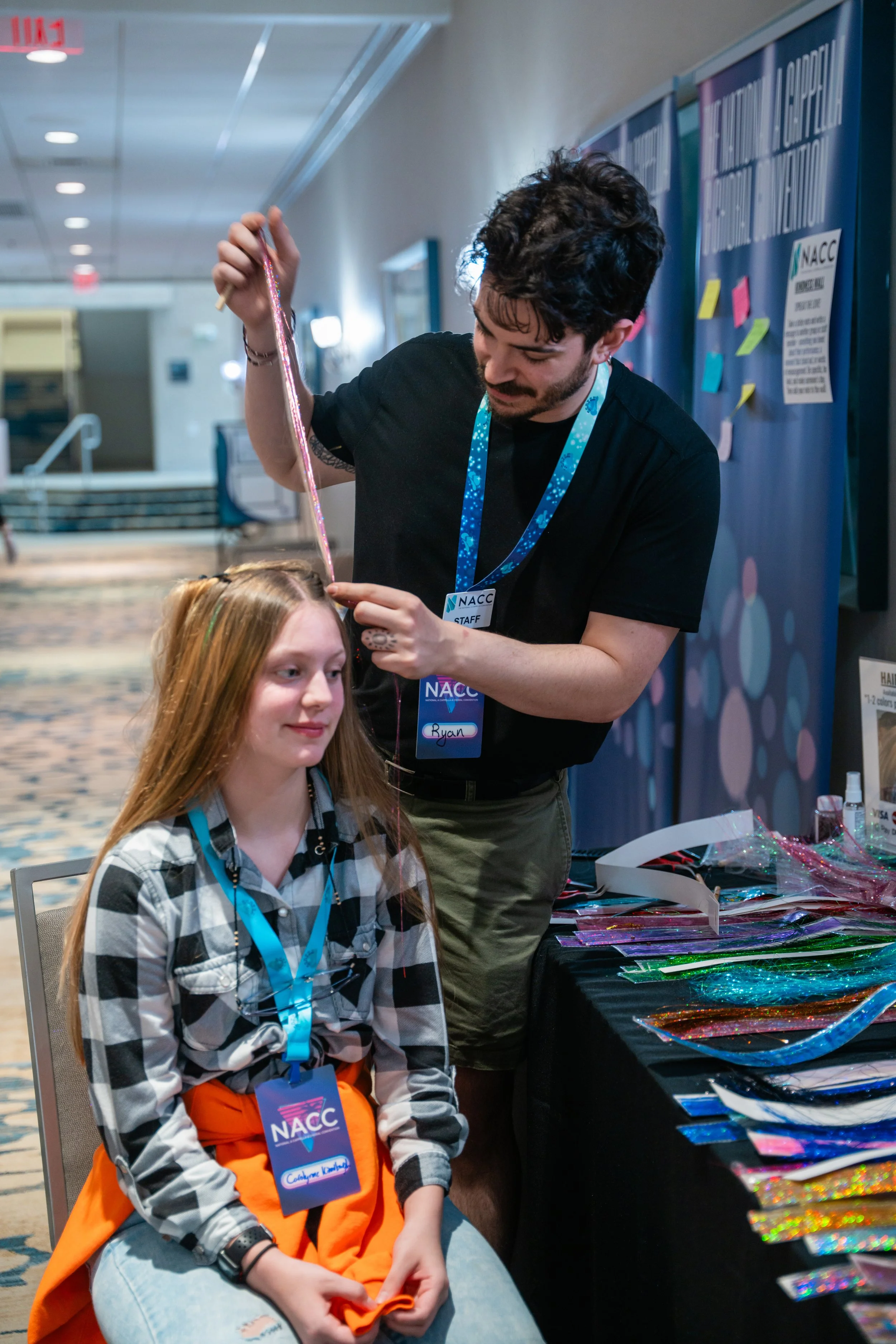 A young girl with long blonde hair, wearing a black-and-white checkered shirt, orange pants, and a blue lanyard, sitting at a table with colorful shiny material. A man with dark hair and a beard, wearing a black shirt, is standing behind her, holding