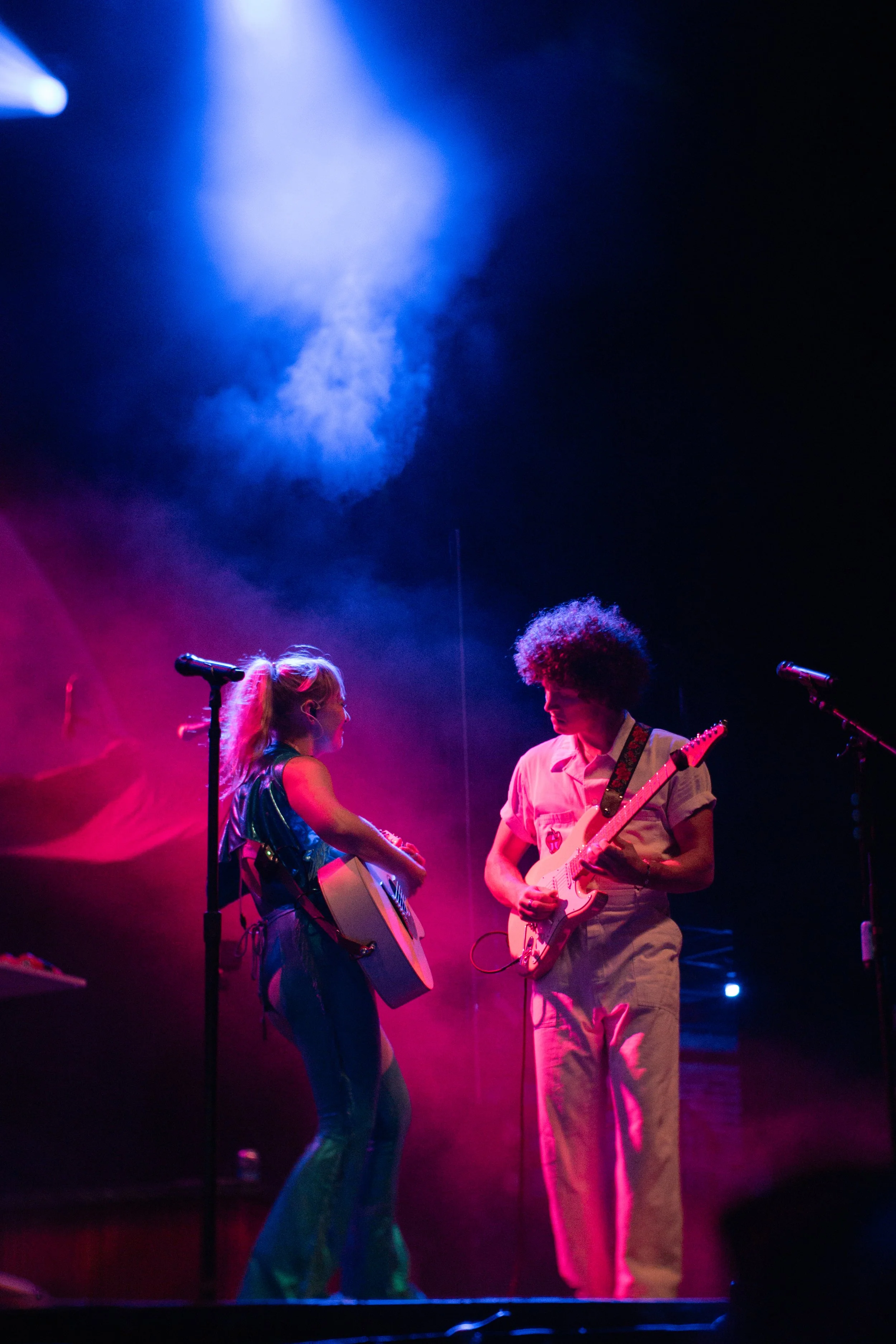 Two musicians performing on stage, one girl with a guitar and a boy with an electric guitar, under colorful stage lights with pink and blue smoke.