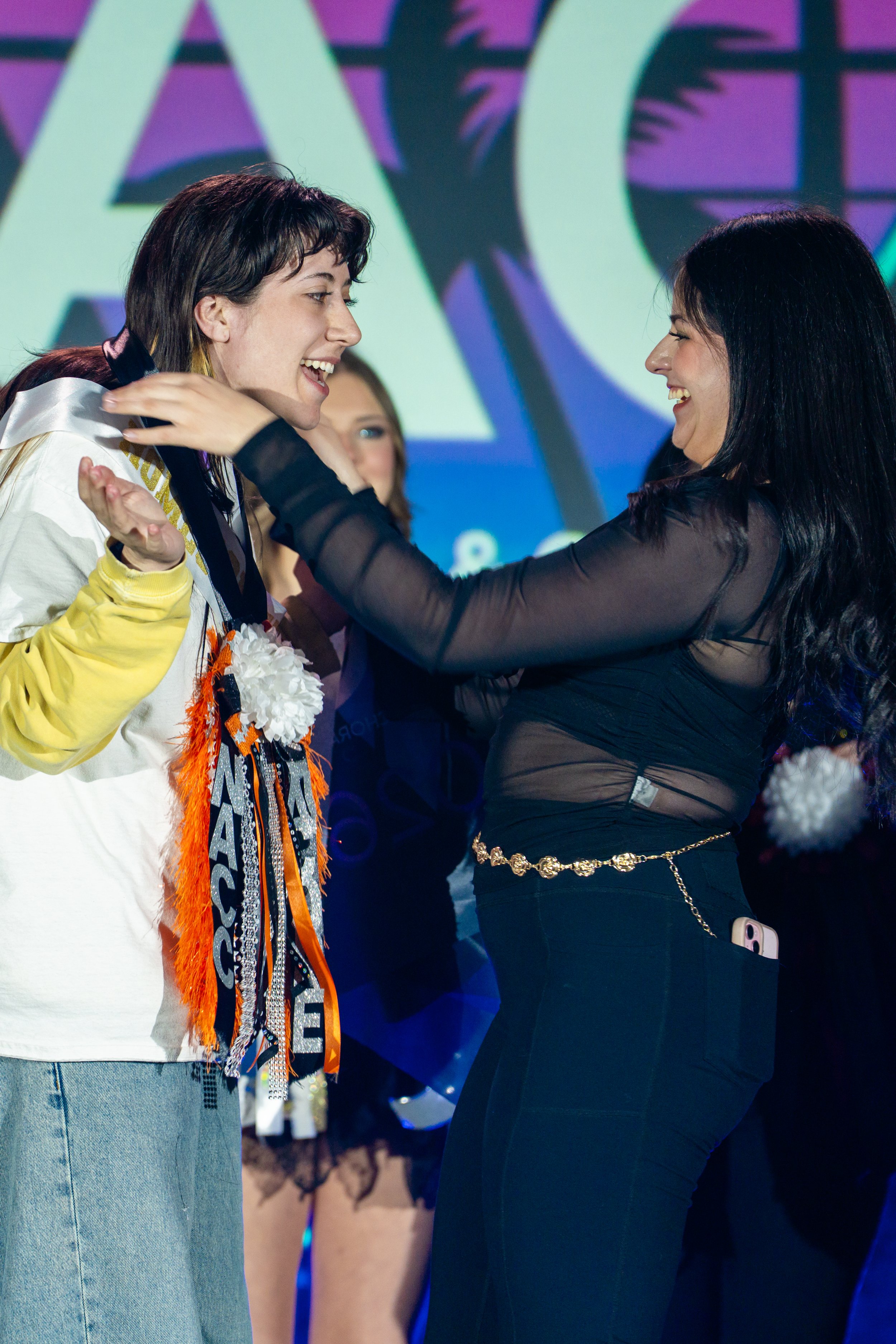 Two women are smiling and talking to each other, facing each other, at an event with a large screen in the background showing graphics. One woman has dark hair and is wearing a black sheer top with a gold chain belt. The other woman has short dark ha