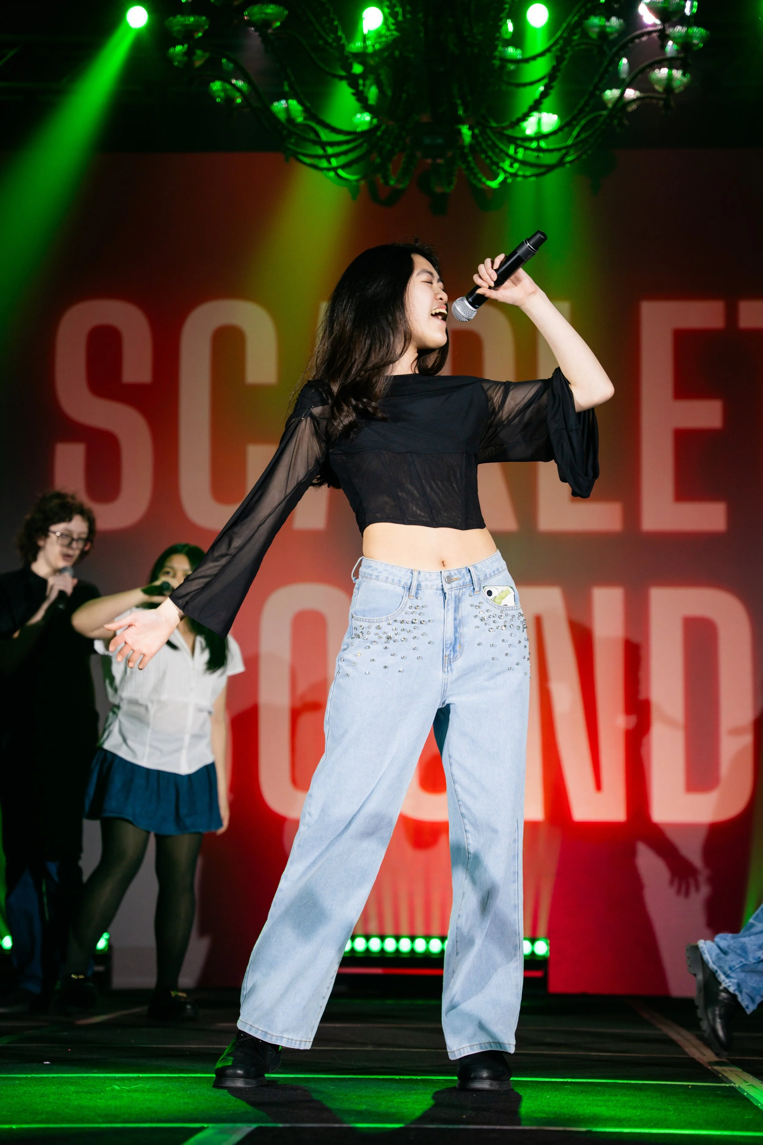 A young woman singing into a microphone on stage with a red background reading 'SOCIAL'. She is wearing a black sheer top and light blue jeans with a phone in her front pocket, and has dark, wavy hair. Green stage lights illuminate the scene, with other people visible in the background.