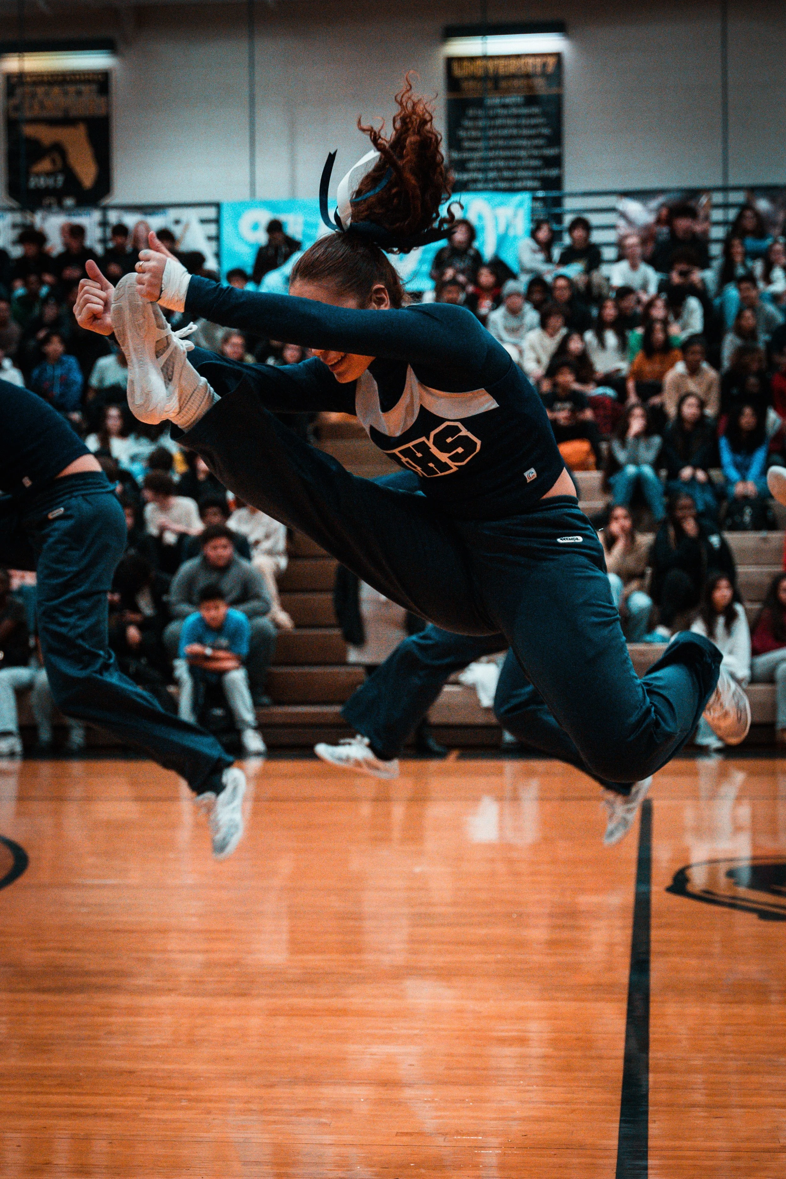 A female cheerleader jumping in mid-air during a cheerleading performance in an indoor gymnasium with a crowd of spectators watching.