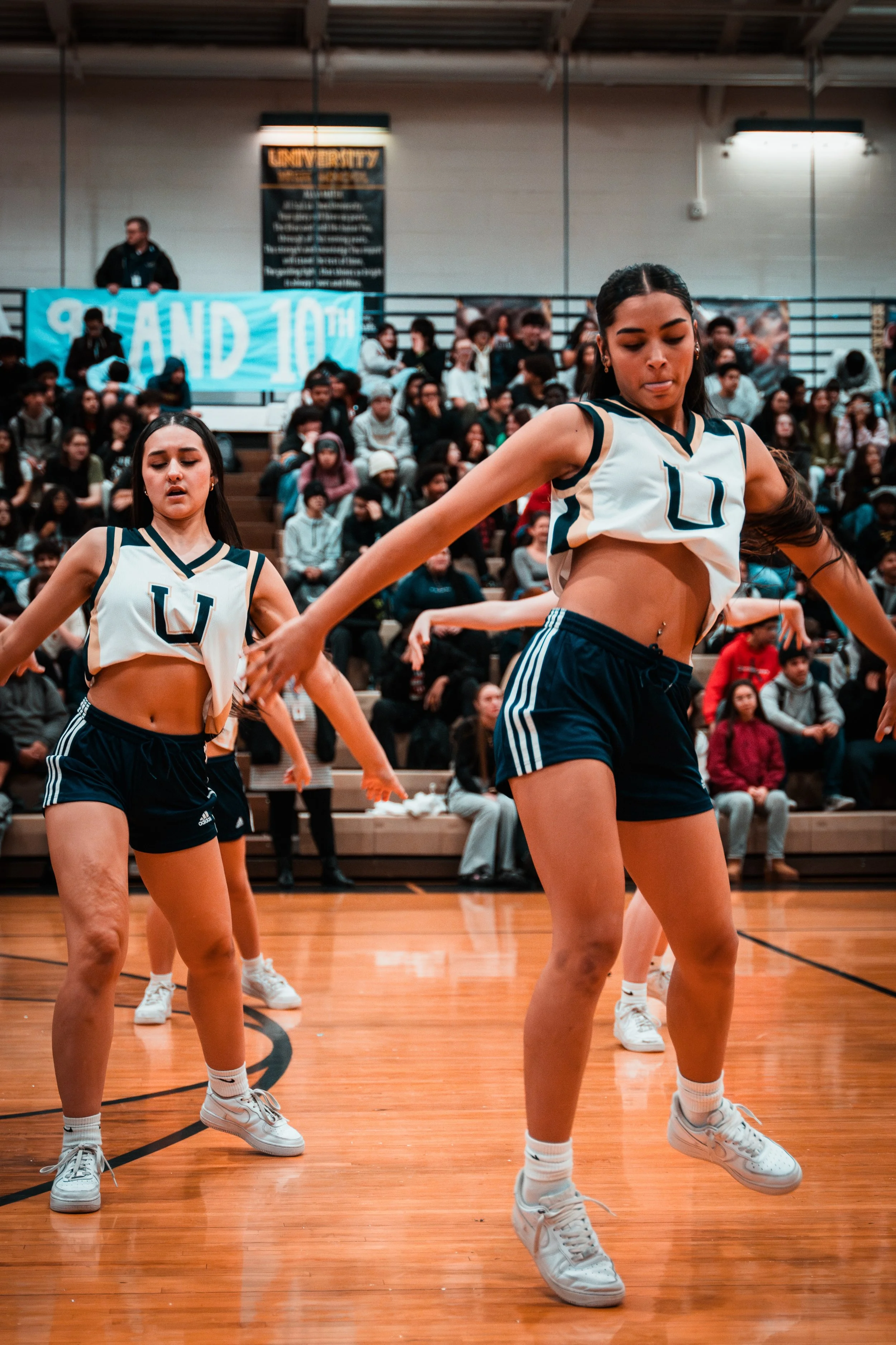 Two female cheerleaders dancing during a basketball game in a gymnasium, with a crowd of spectators in the background.