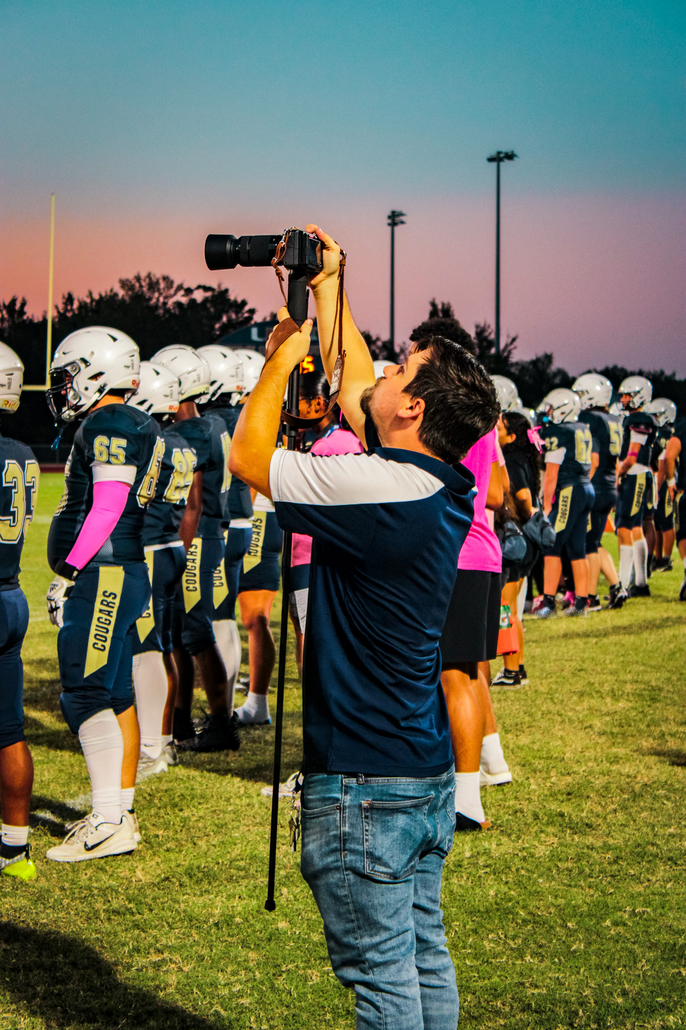 A man filming with a camera on a football field during sunset, with football players in uniforms lined up in the background.