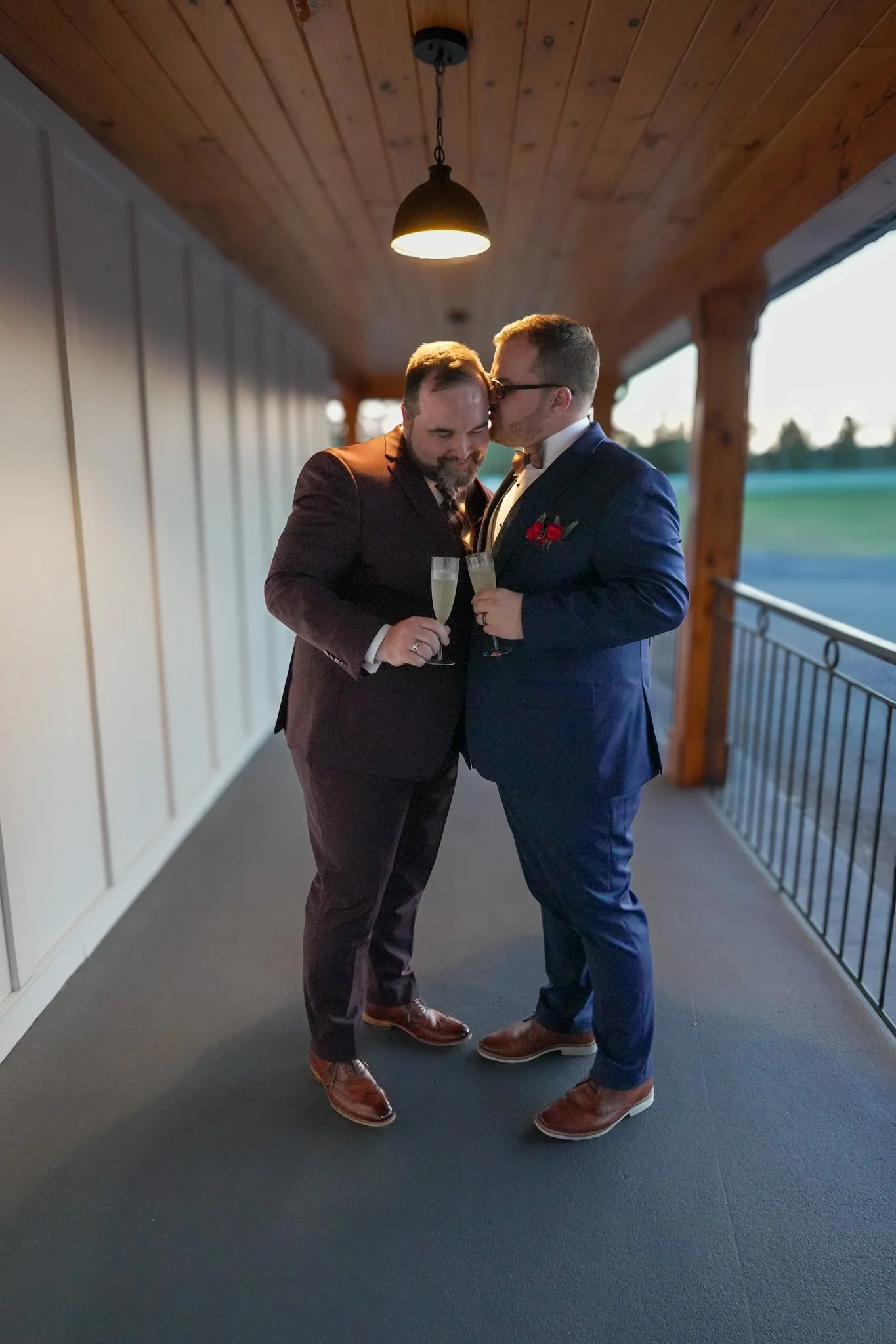 Two men in suits sharing a kiss while holding champagne glasses on a balcony with wood ceiling, white wall, and sunset scenery in the background.
