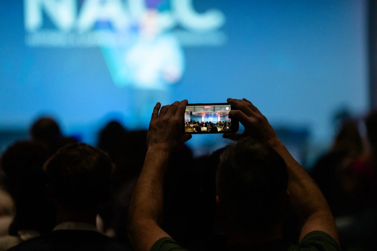 Person taking a photo with a smartphone at a conference or event, with a large screen displaying a blue background and text in the background.