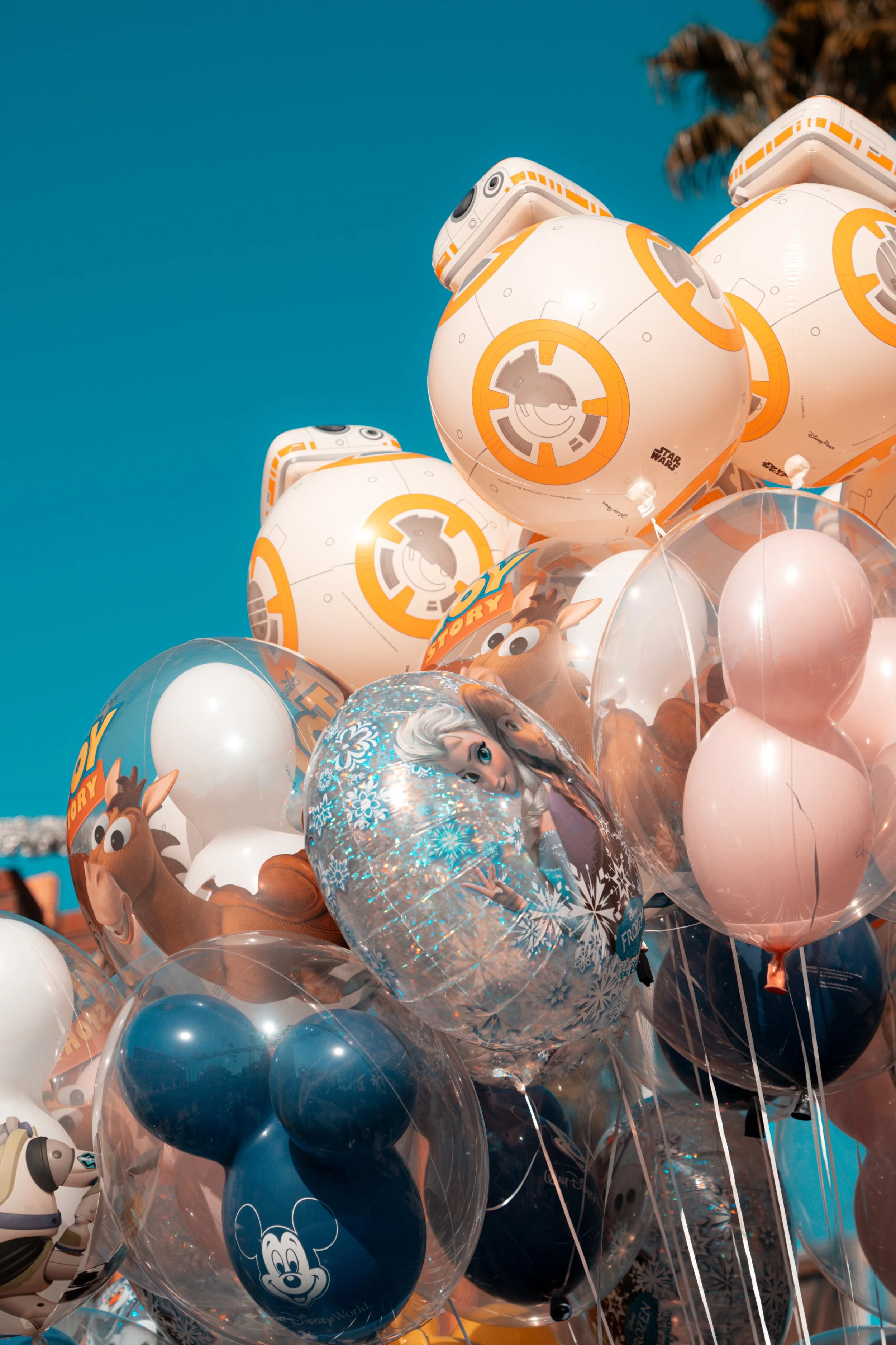 A cluster of Disney and Star Wars themed balloons, including characters like Elsa from Frozen, Mickey Mouse, and BB-8 from Star Wars, against a clear blue sky.