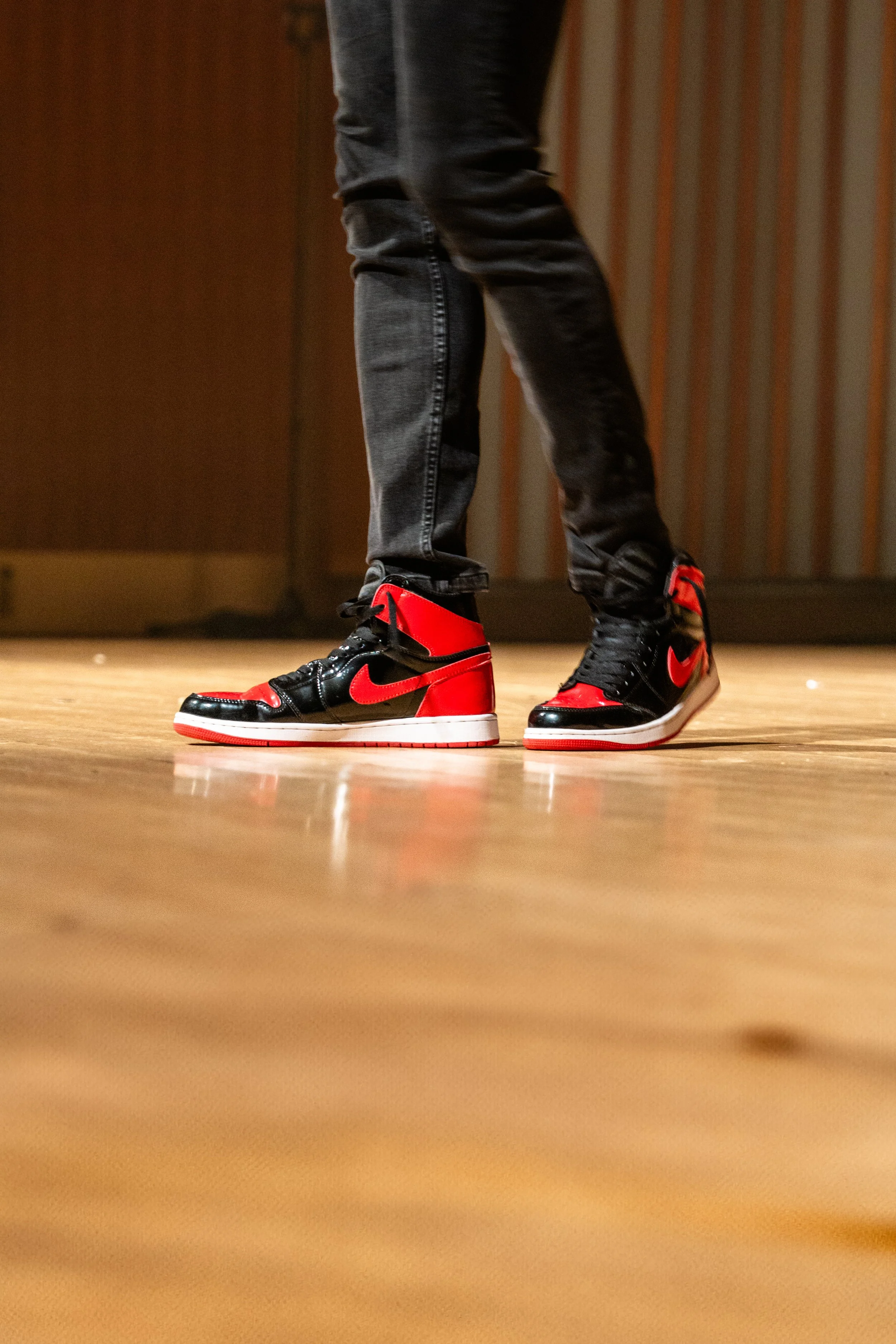 Person wearing black jeans and red and black Nike sneakers standing on wooden floor.