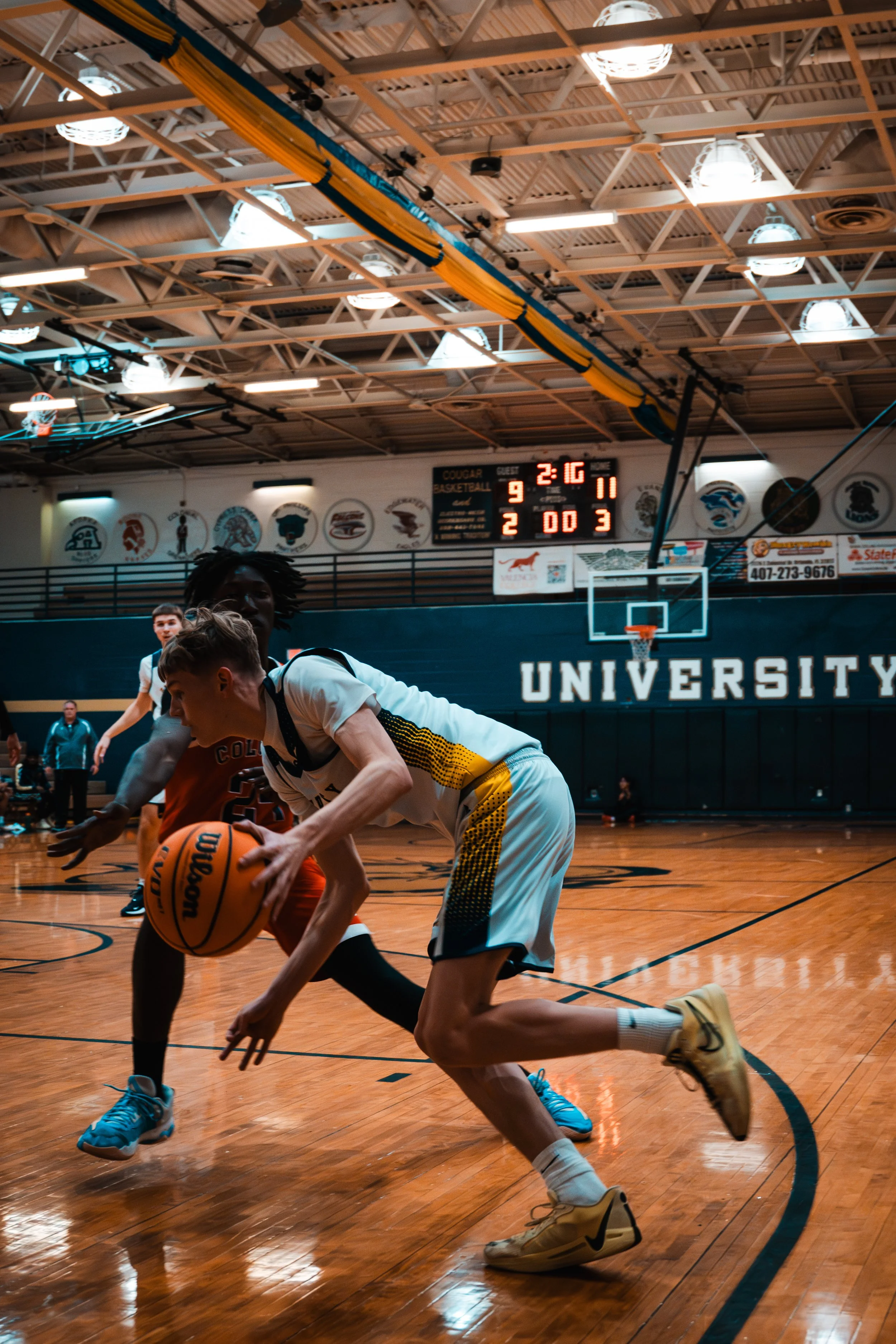 Two basketball players compete for the ball on a wooden court during a game at a university gymnasium. The scoreboard shows 2:16 remaining in the game, with the visiting team leading 9-2.