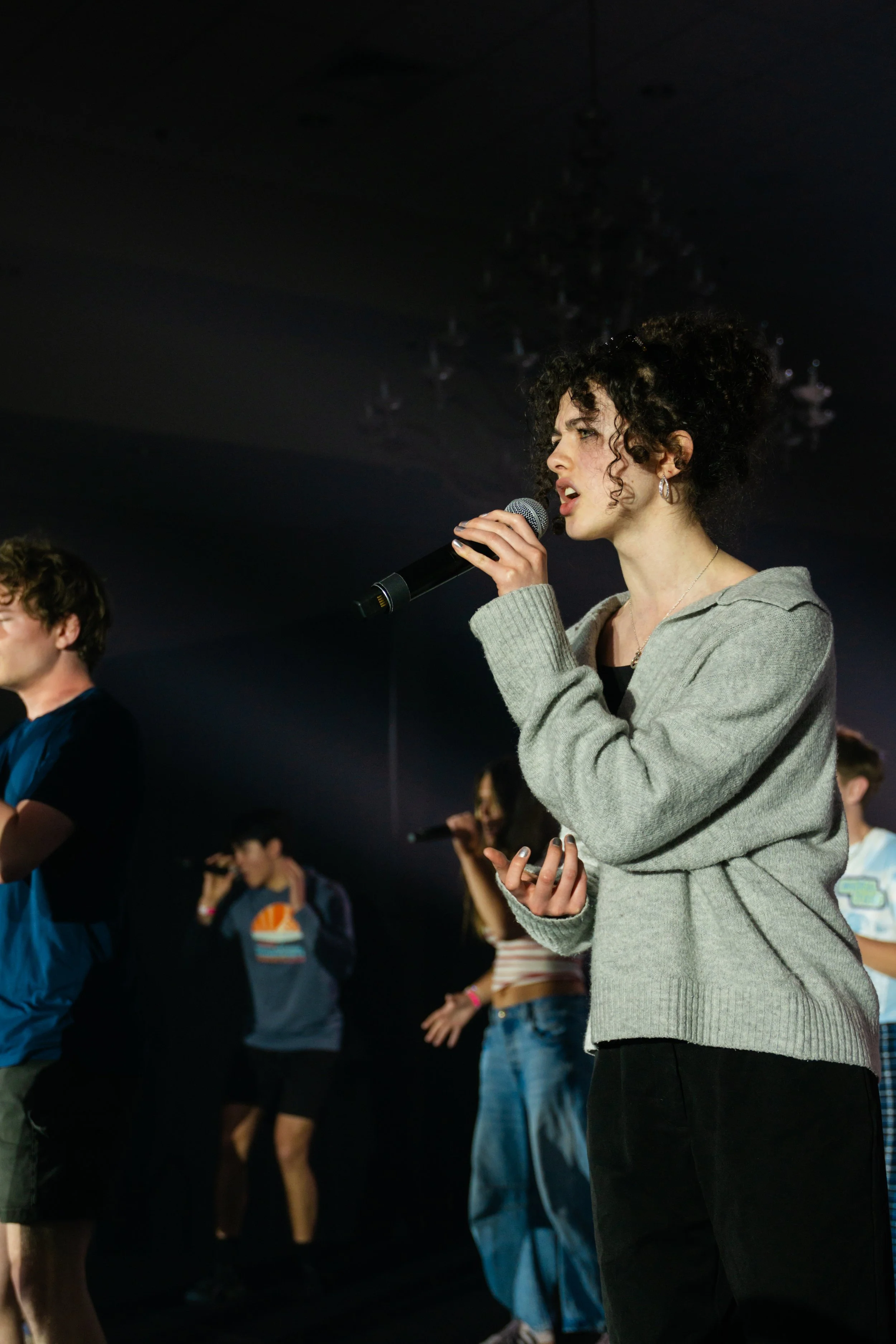 A woman with curly dark hair holds a microphone and appears to be speaking at a gathering or event with multiple people in the background.