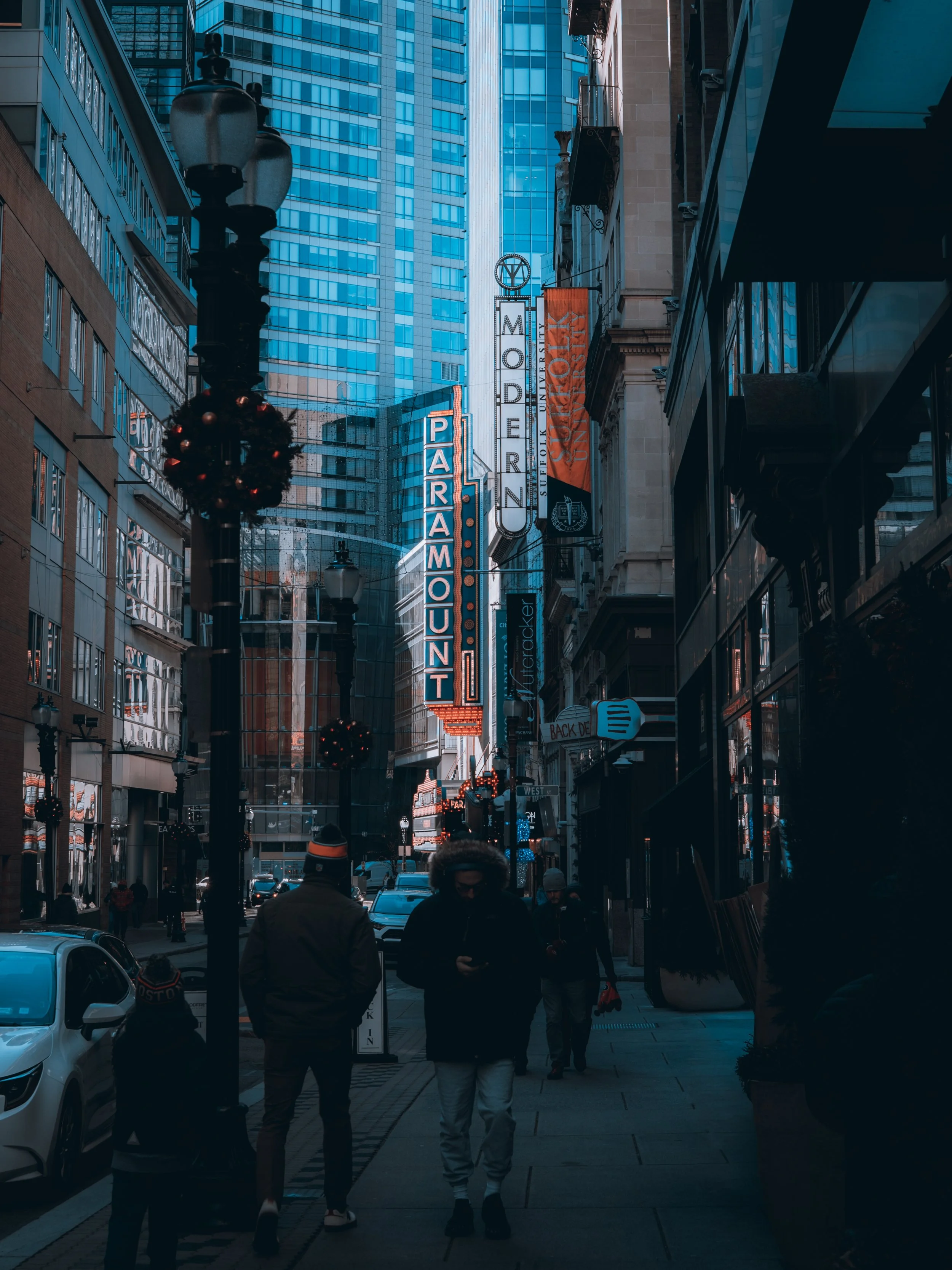 City street scene with pedestrians walking, tall buildings, neon signs, and Christmas decorations on lampposts.