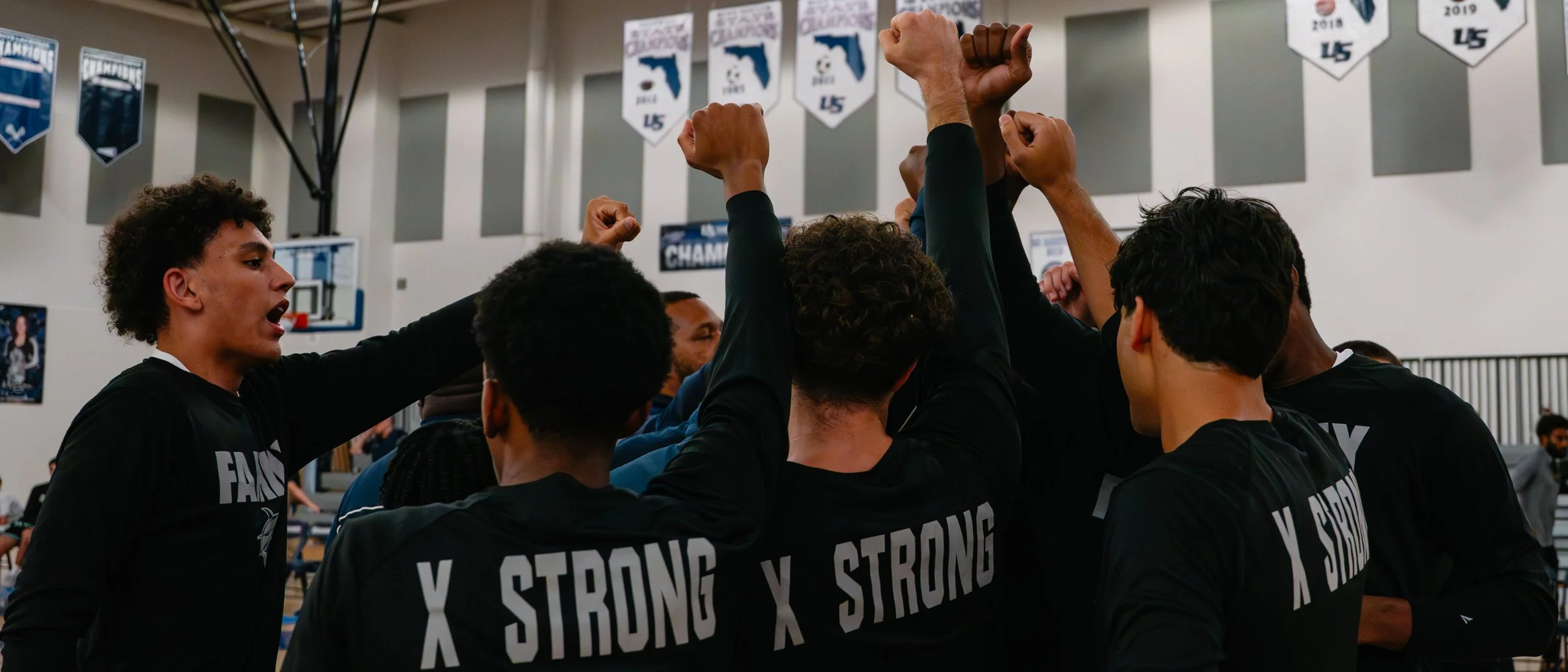 A group of basketball players wearing black jerseys with 'X STRONG' on the back, cheering and raising their fists together in a gymnasium with banners hanging from the ceiling.