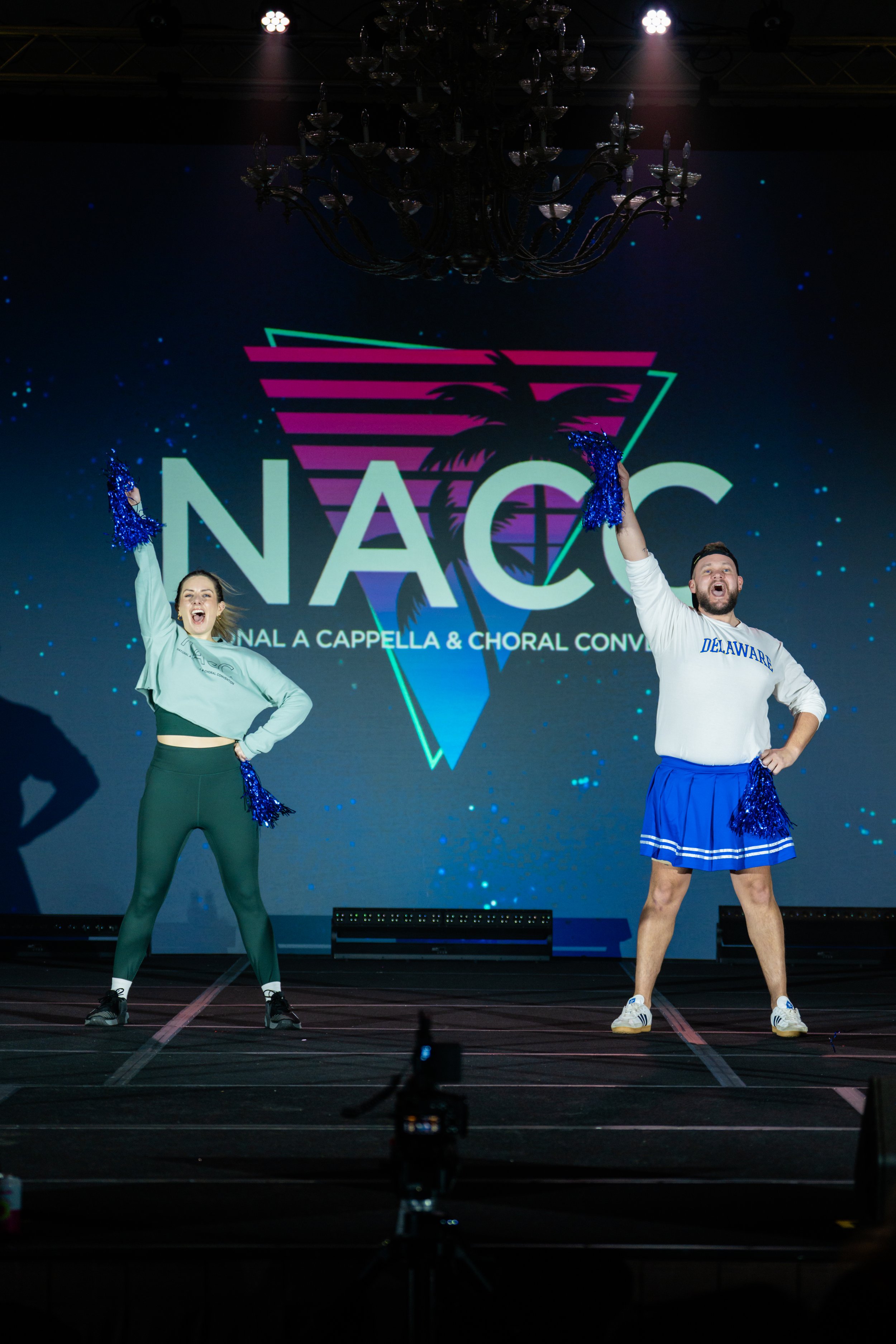 Two cheerleaders performing on stage with a large screen behind them displaying the NAC logo and palm trees. One cheerleader is wearing green leggings and a white top, the other is wearing a blue skirt and a white sweatshirt with 'Delaware' written o